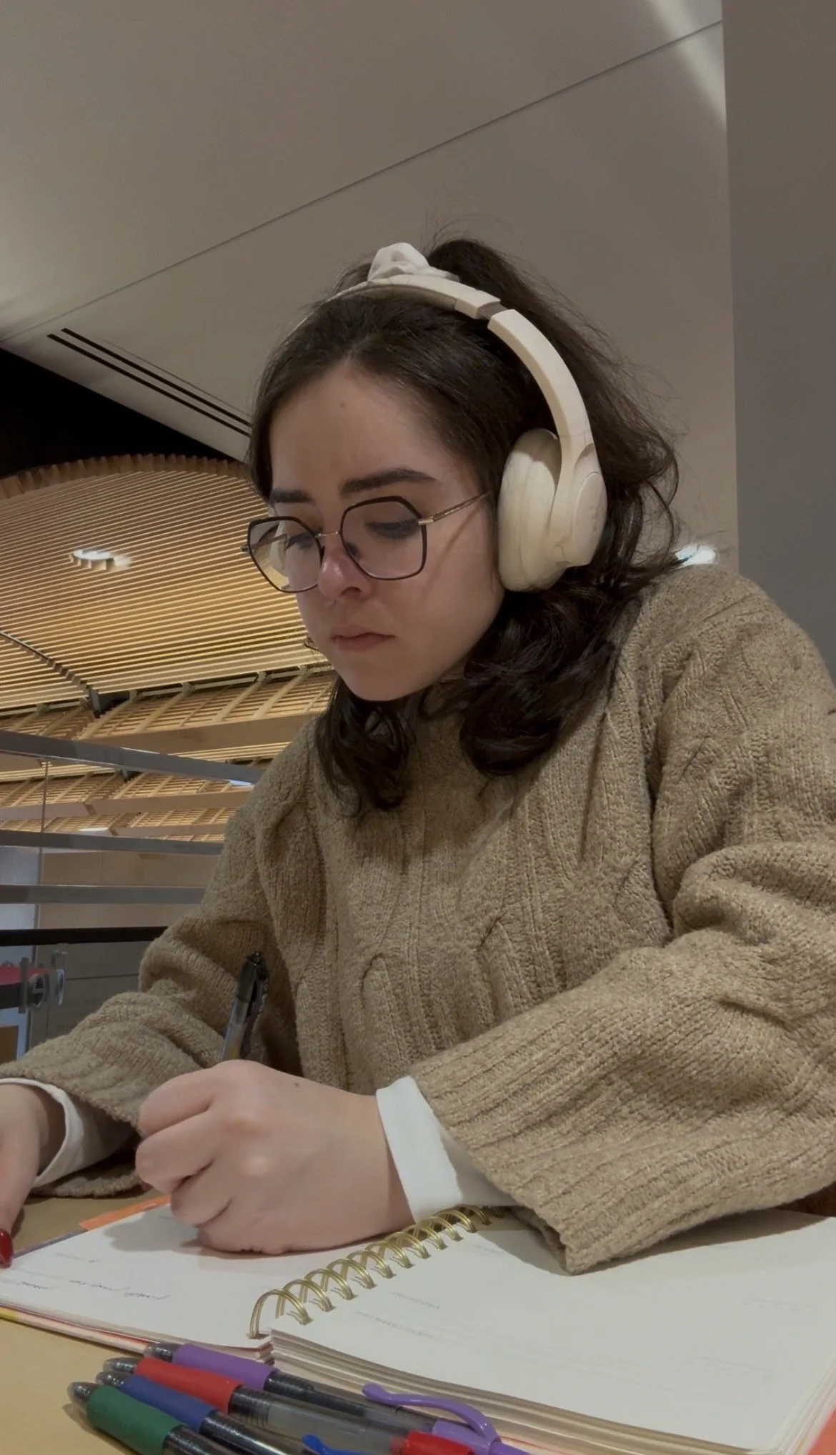 A young woman with glasses and dark wavy hair is sitting at a table, writing in a notebook, with multiple colorful pens nearby. She is wearing headphones and a beige sweater, and appears to be studying or working.