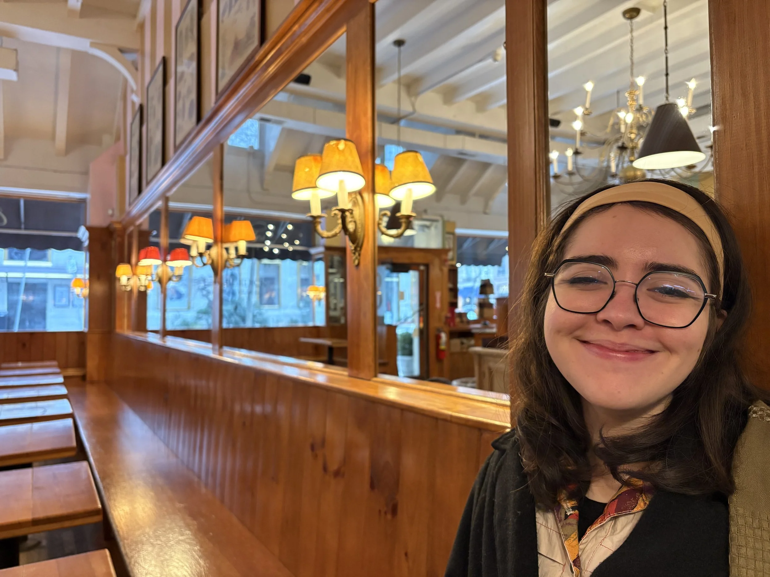 A young woman with glasses and a headband smiling inside a cozy, warmly lit restaurant with wooden walls, framed pictures, and chandeliers.
