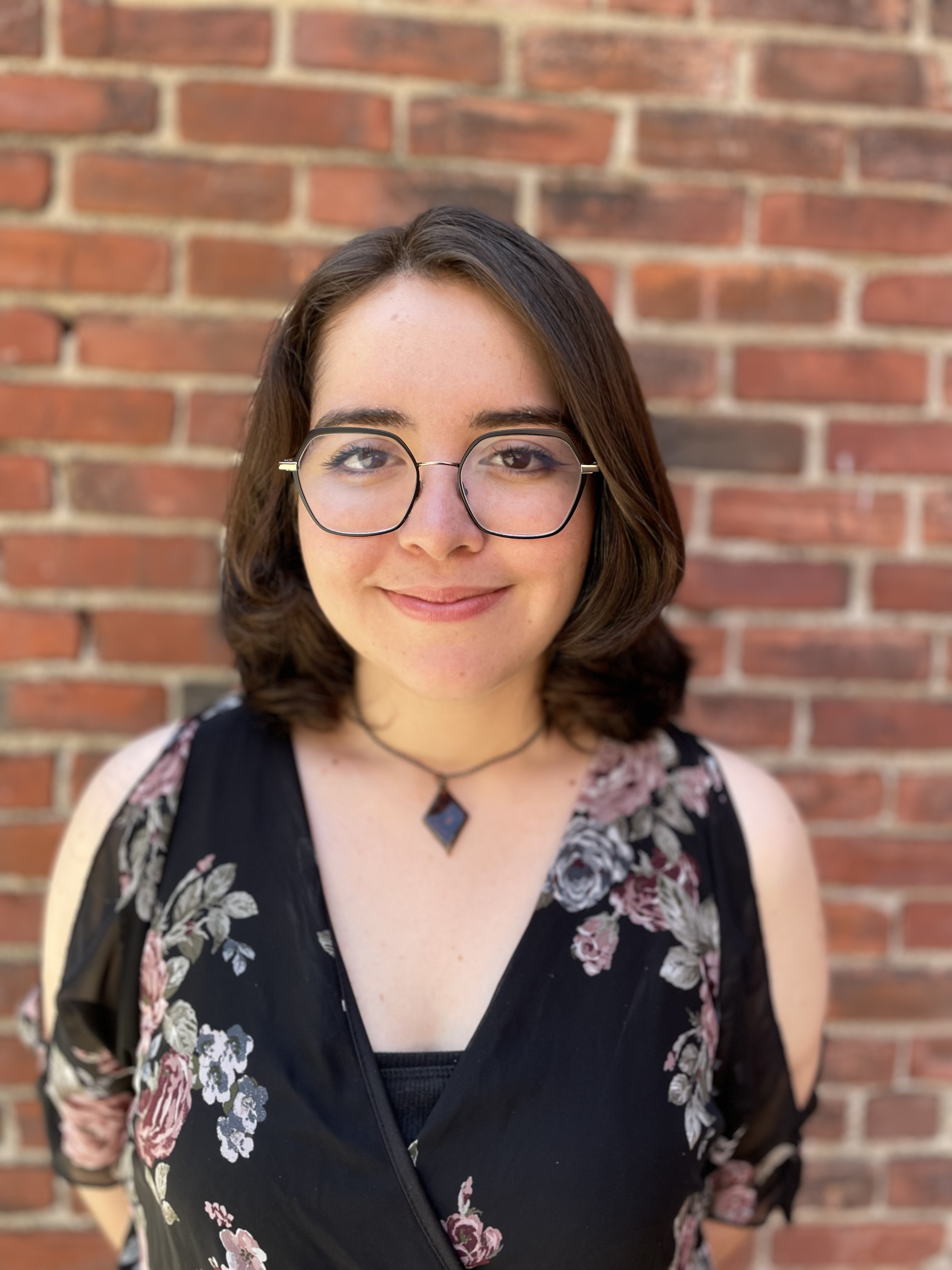 A young woman with shoulder-length brown hair, glasses, and a black floral dress standing in front of a red brick wall.