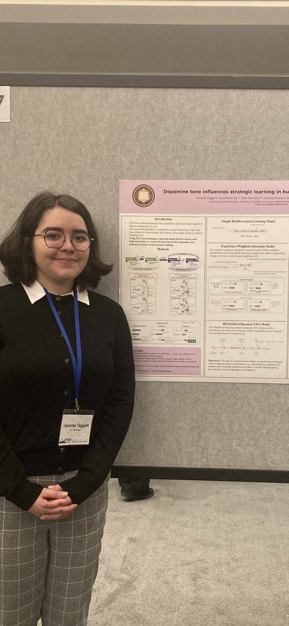 A young woman wearing glasses and a black cardigan with a white collared shirt, standing in front of a scientific research poster at a conference. She has a name tag that reads 'Jacinda Taggert' and is smiling.