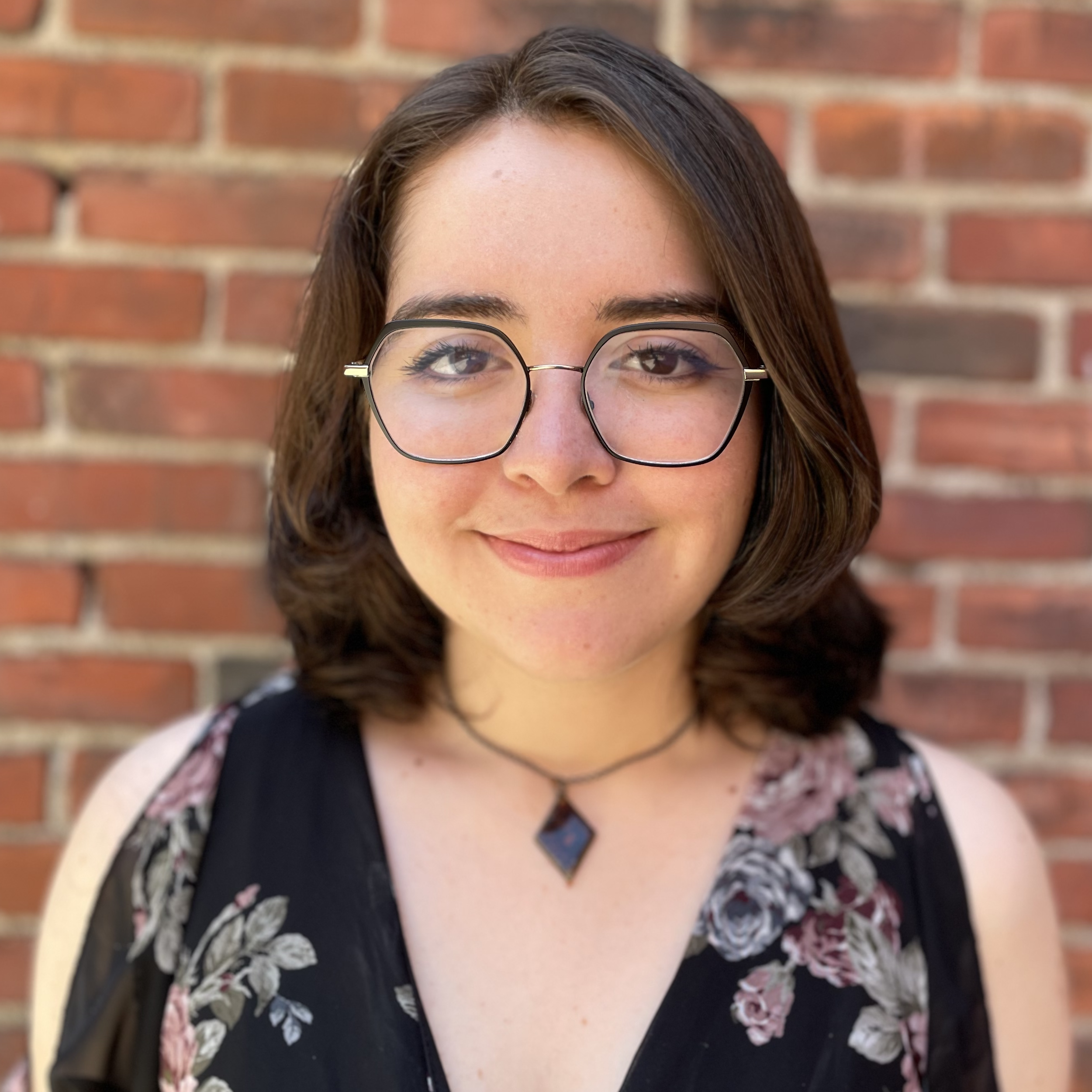 Close-up of a woman with brown hair, glasses, and a floral dress standing in front of a brick wall.