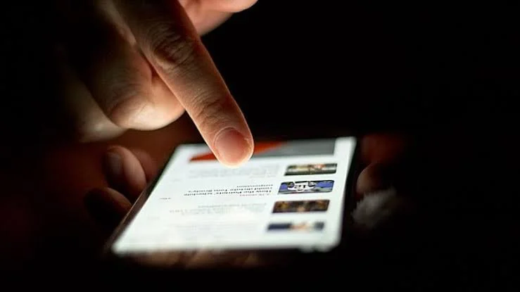 A person taps on a smartphone screen in a dark environment, with the screen displaying a webpage or app.