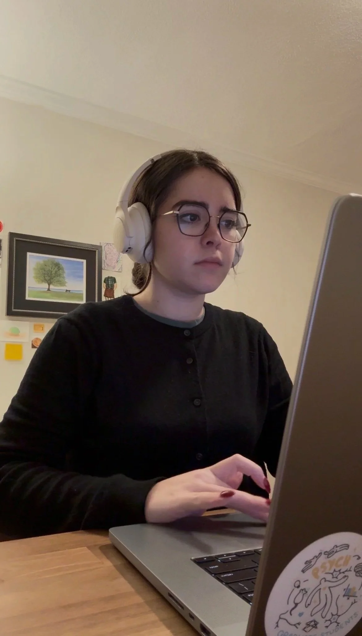 A young woman with dark hair, wearing glasses and headphones, working on a laptop at a wooden table in a room with wall art.