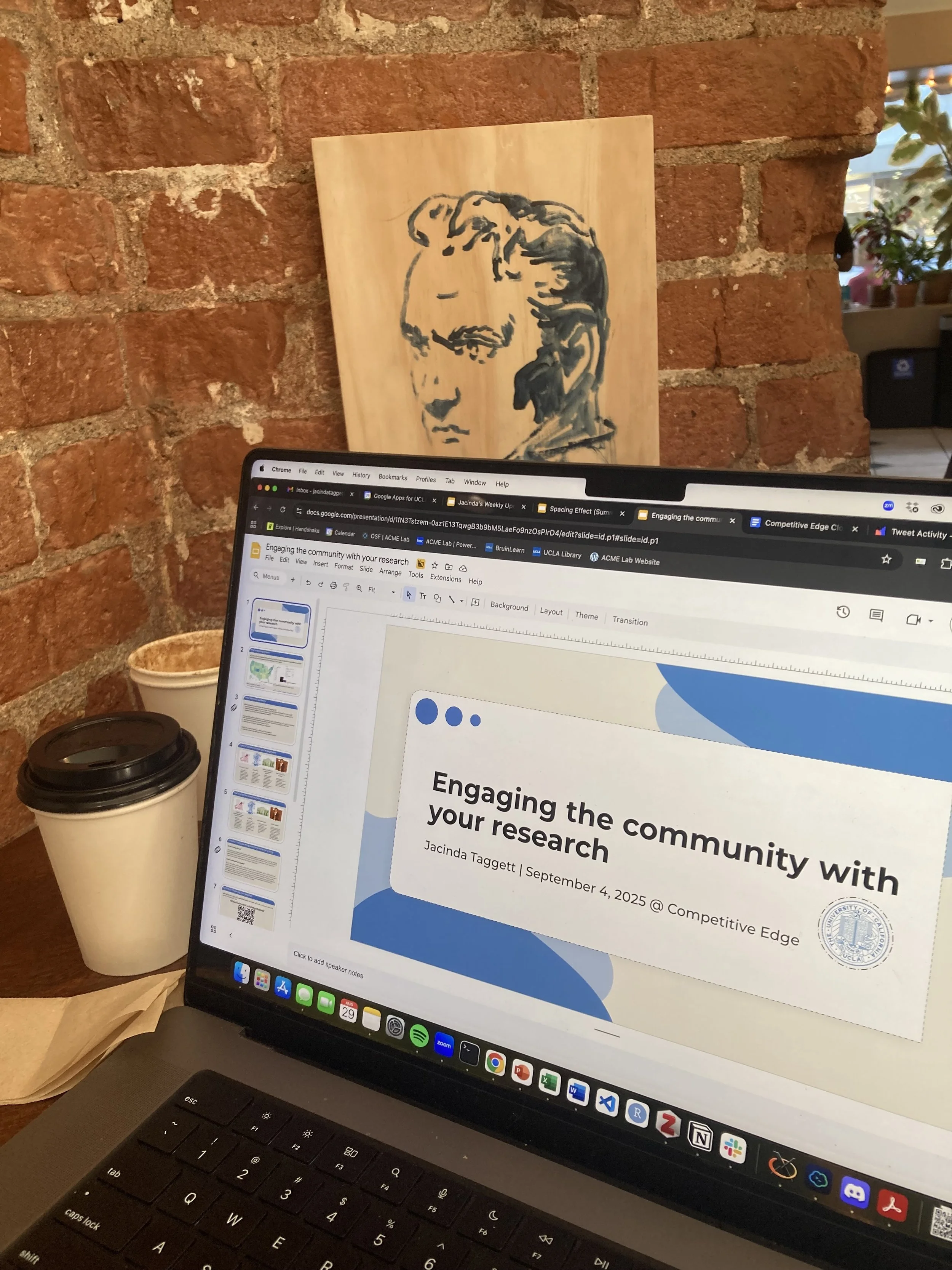 A computer screen displaying a presentation slide titled 'Engaging the community with your research' by Jacinda Taggett. There is a brick wall in the background with a wooden artwork of a person’s profile, and a paper coffee cup beside the laptop.