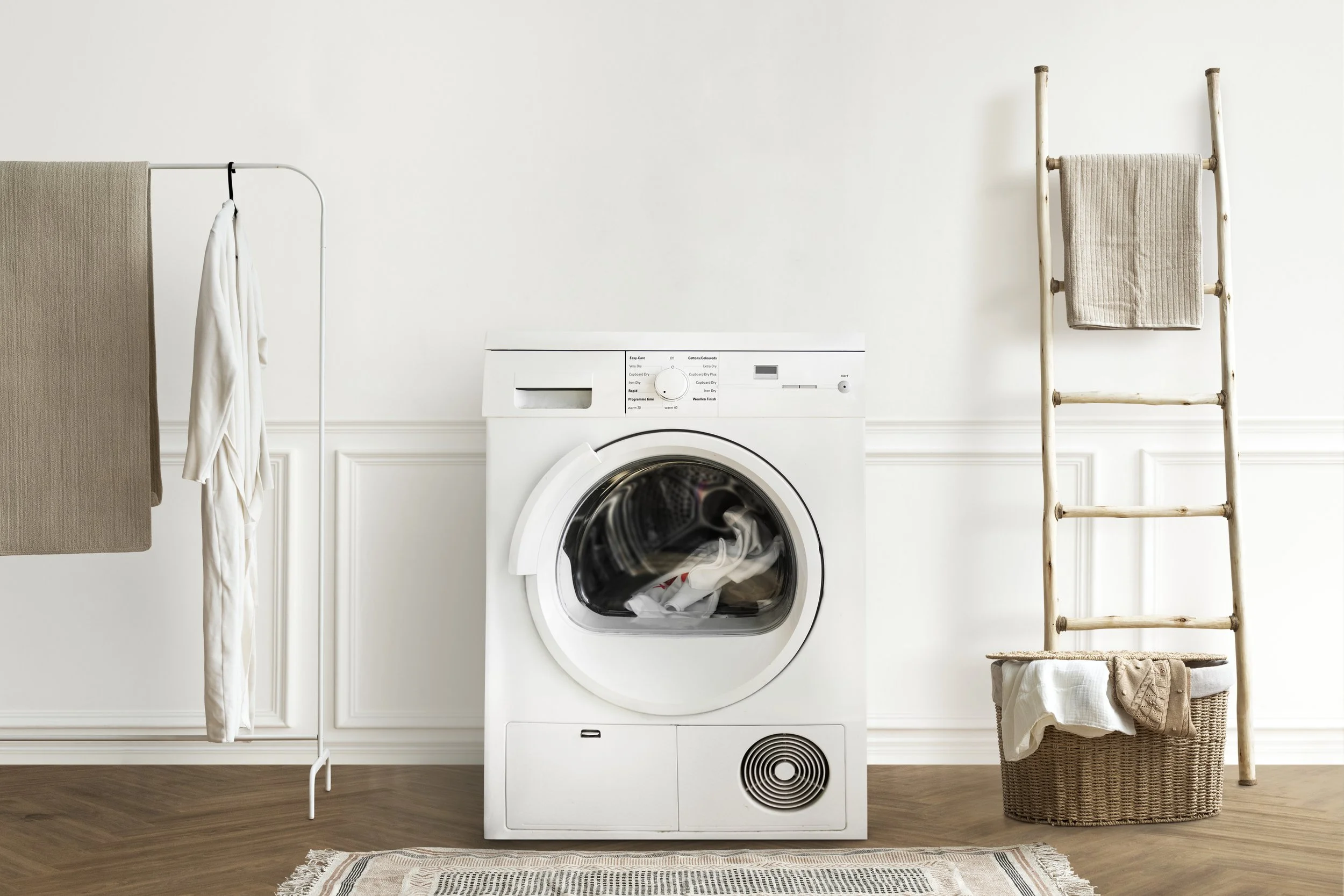A laundry room with a white washing machine, a wooden ladder with a beige towel, a laundry basket with clothes, and a drying rack with towels, all on a wooden floor with a beige rug.