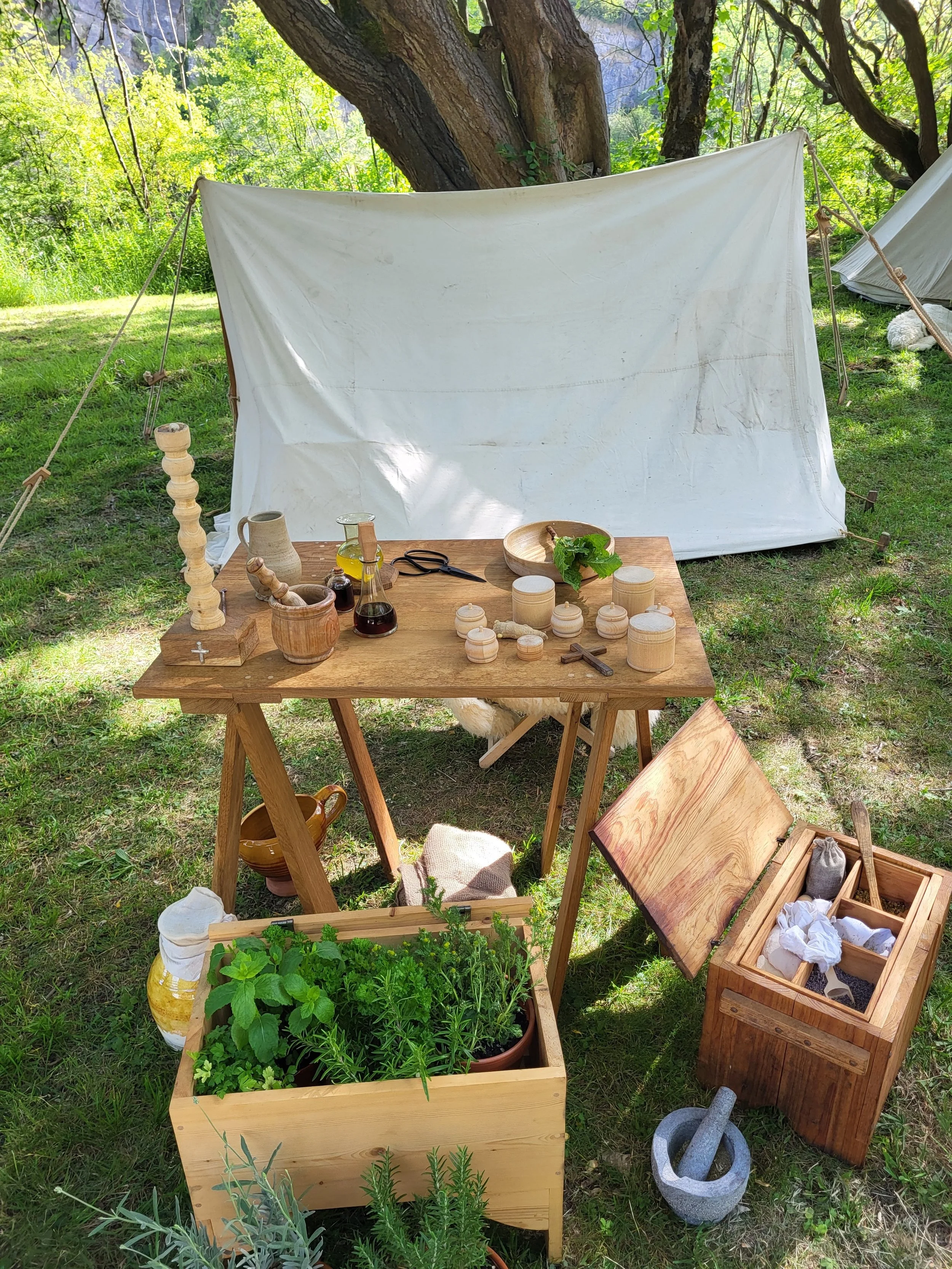 A display of gardening and woodworking tools outdoors, including a table with jars, scissors, and plants, a wooden box with herbs, and a trellis made of wooden slats. Behind the table, there is a white fabric or canvas backdrop attached to trees in a lush green outdoor setting.