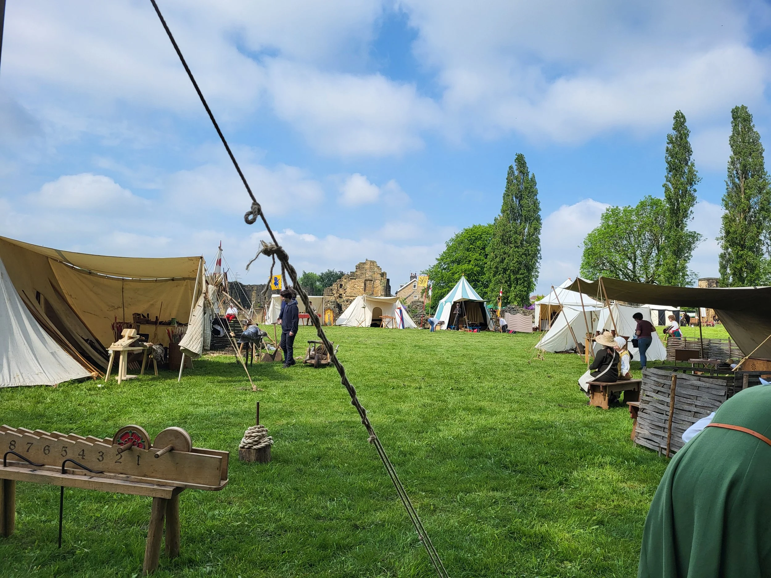 A historical reenactment event with tents, banners, and people in period clothing outdoors on a sunny day with green grass and tall trees.