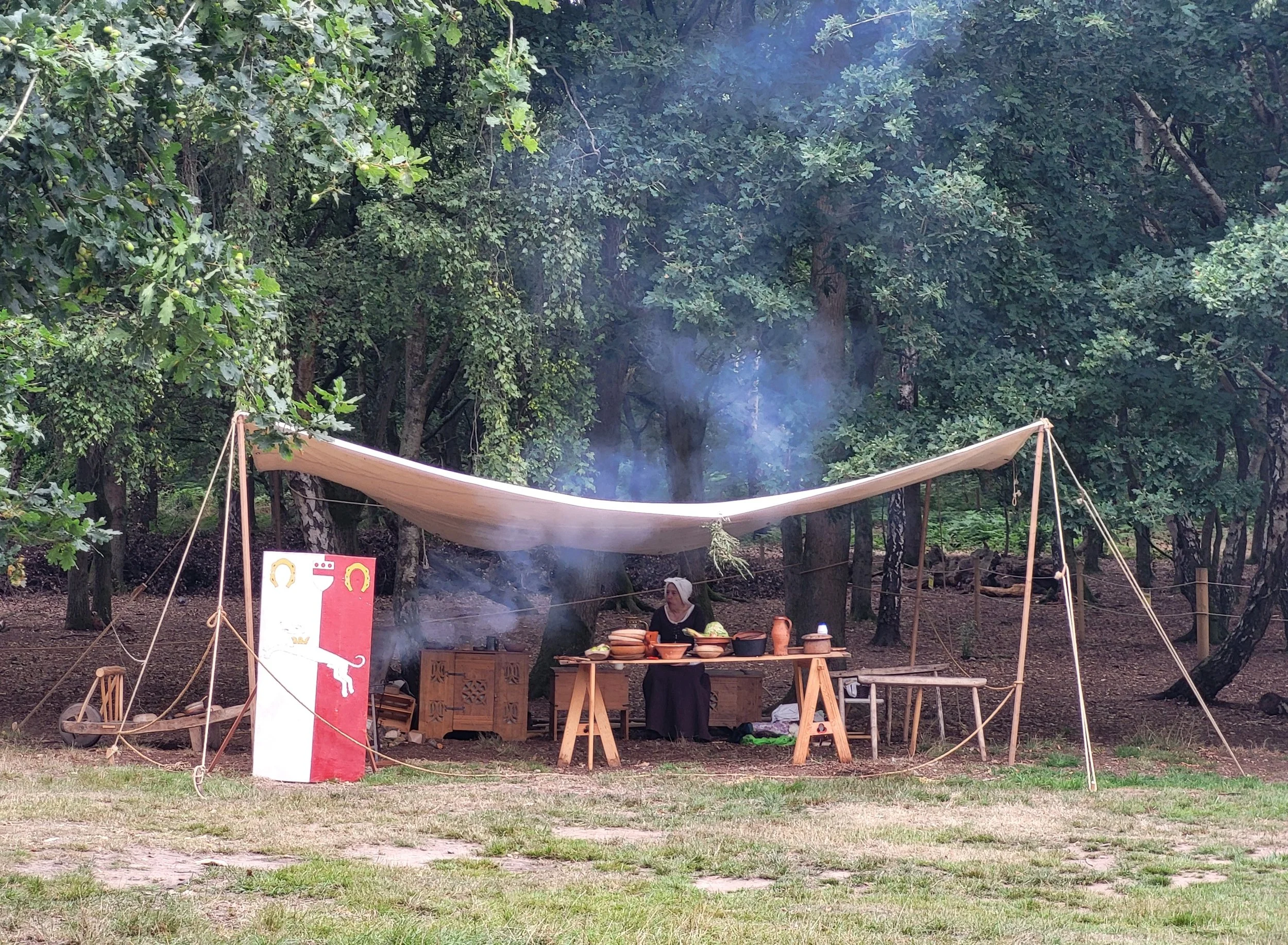 A medieval-themed outdoor market stall in a wooded area, with a pavilion and a woman sitting behind a table with pottery and food. There is a red and white sign with a crown and animal silhouette, and a small shield with a red cross.