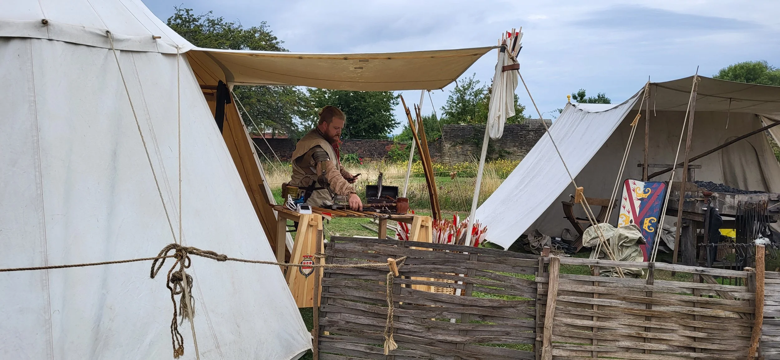 A man dressed in medieval or renaissance costume working at a crafting table set up in a historical reenactment campsite with tents and medieval weapons, armor, and shields around him.
