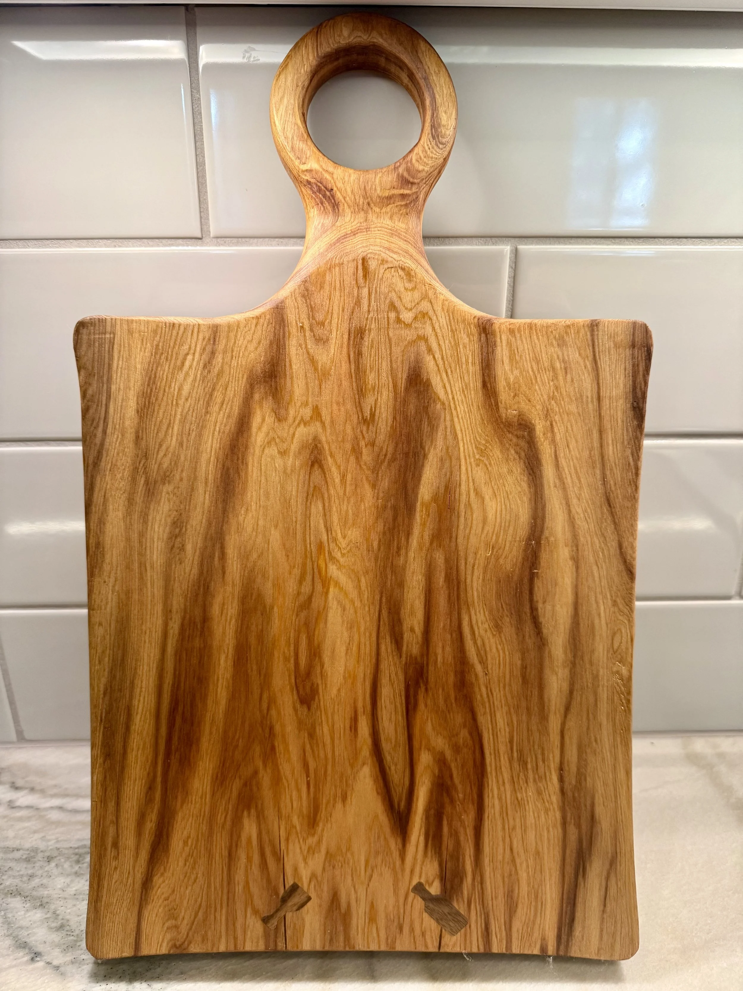 A rectangular wooden cutting board with a round handle at the top, placed against a tiled wall.