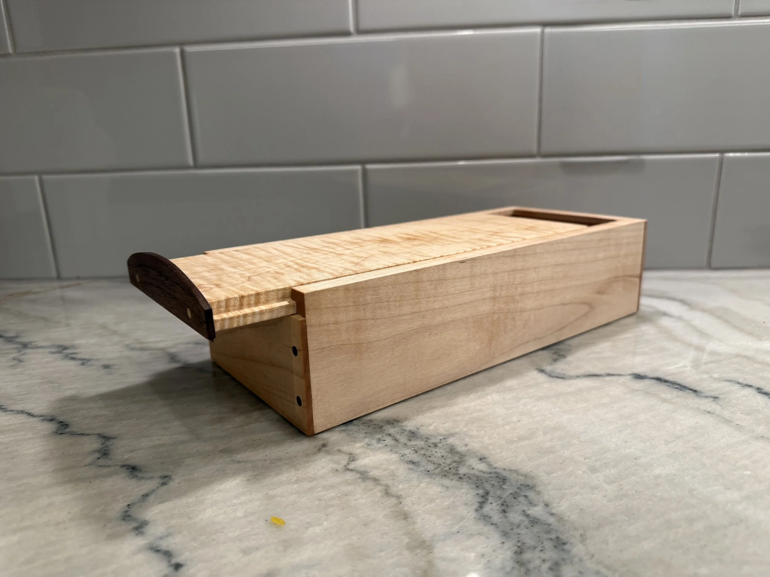 A wooden tissue box holder with a darker wood handle on a marble countertop, gray tiled wall in the background.
