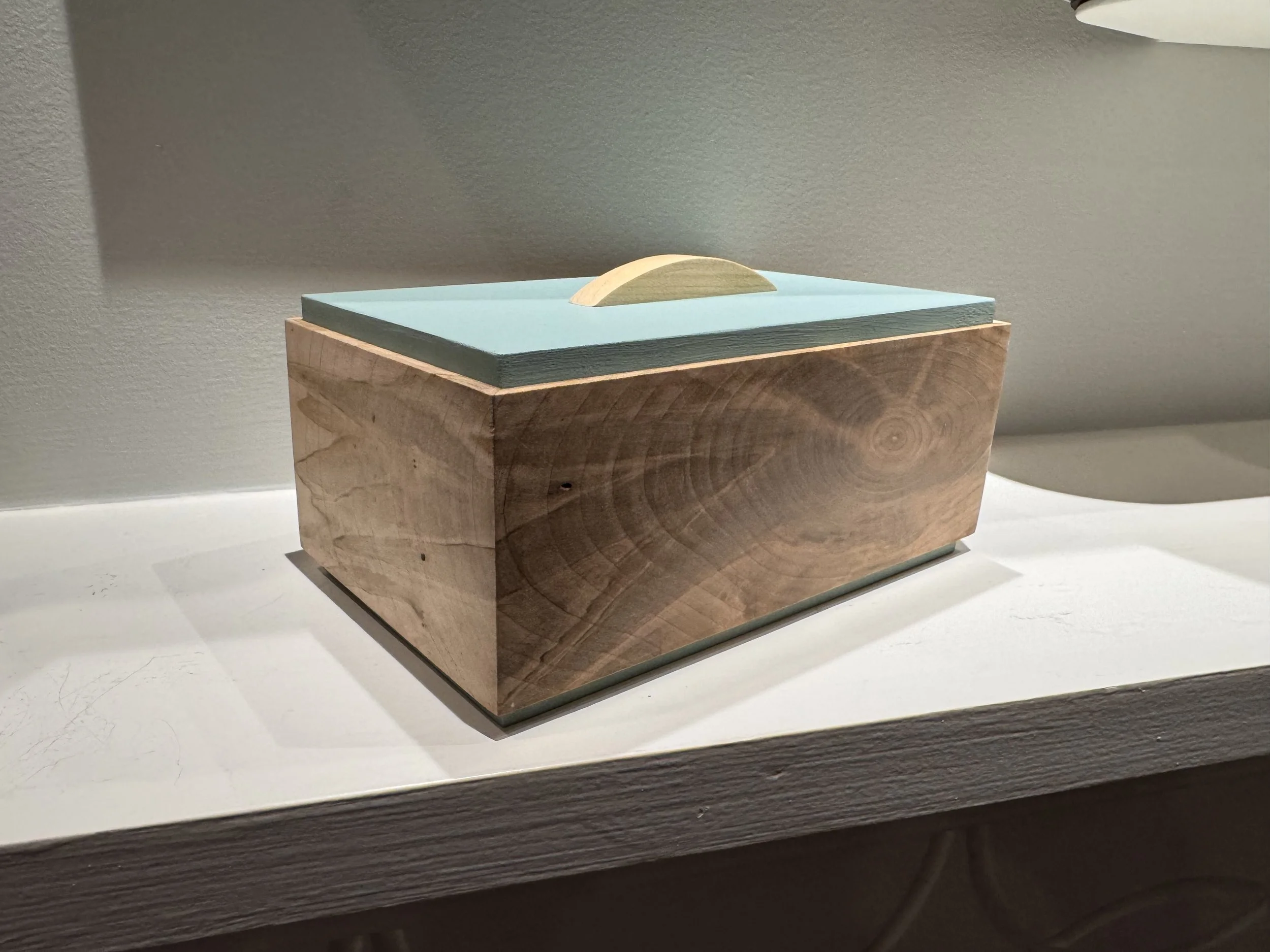 A wooden tissue box with a light blue lid and a beige handle on top, sitting on a white surface against a plain wall.