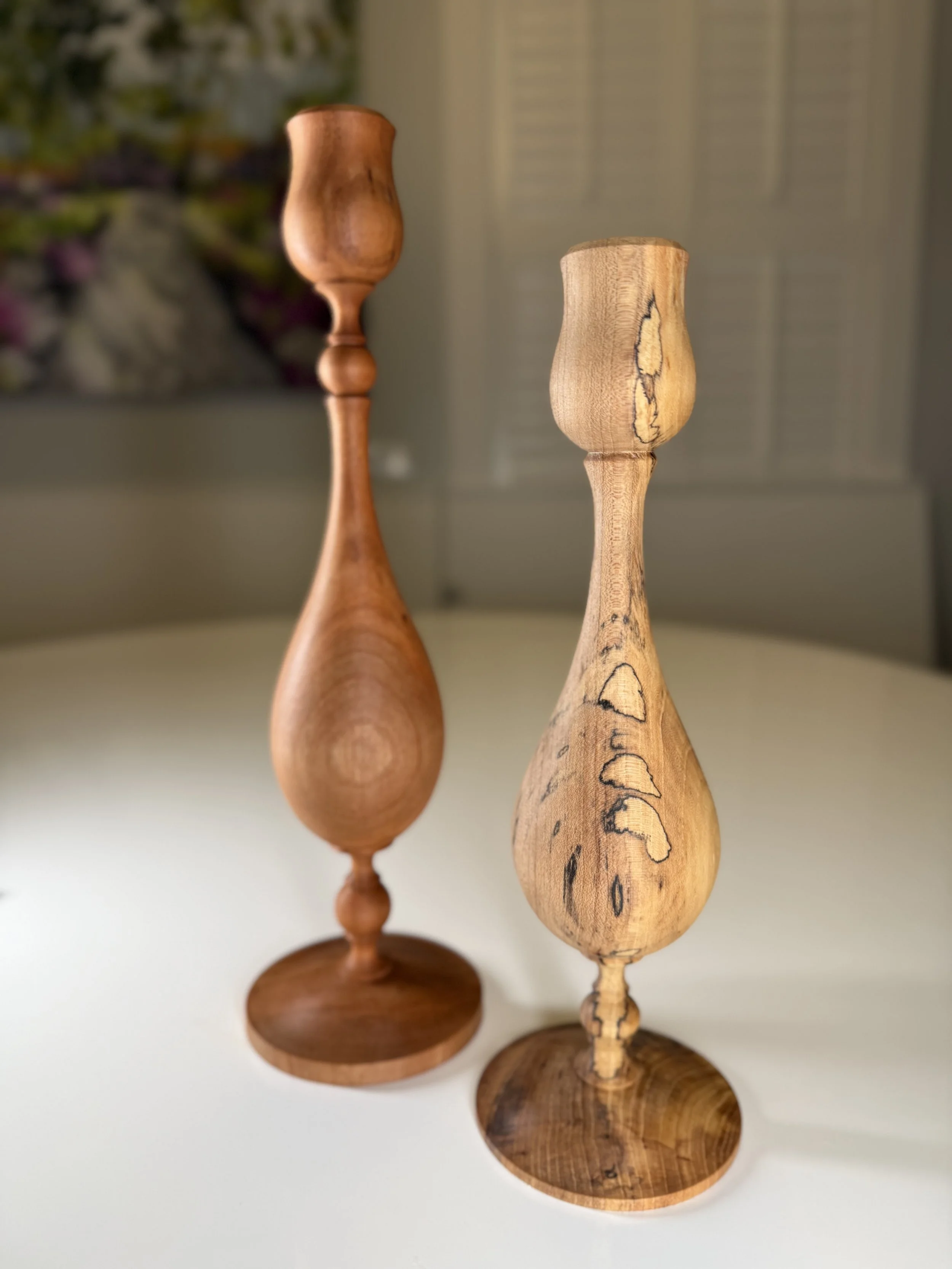Two wooden candlestick holders with bulbous stems, one with a smooth finish and the other with a natural, distressed look, sit on a white surface against a blurred background.