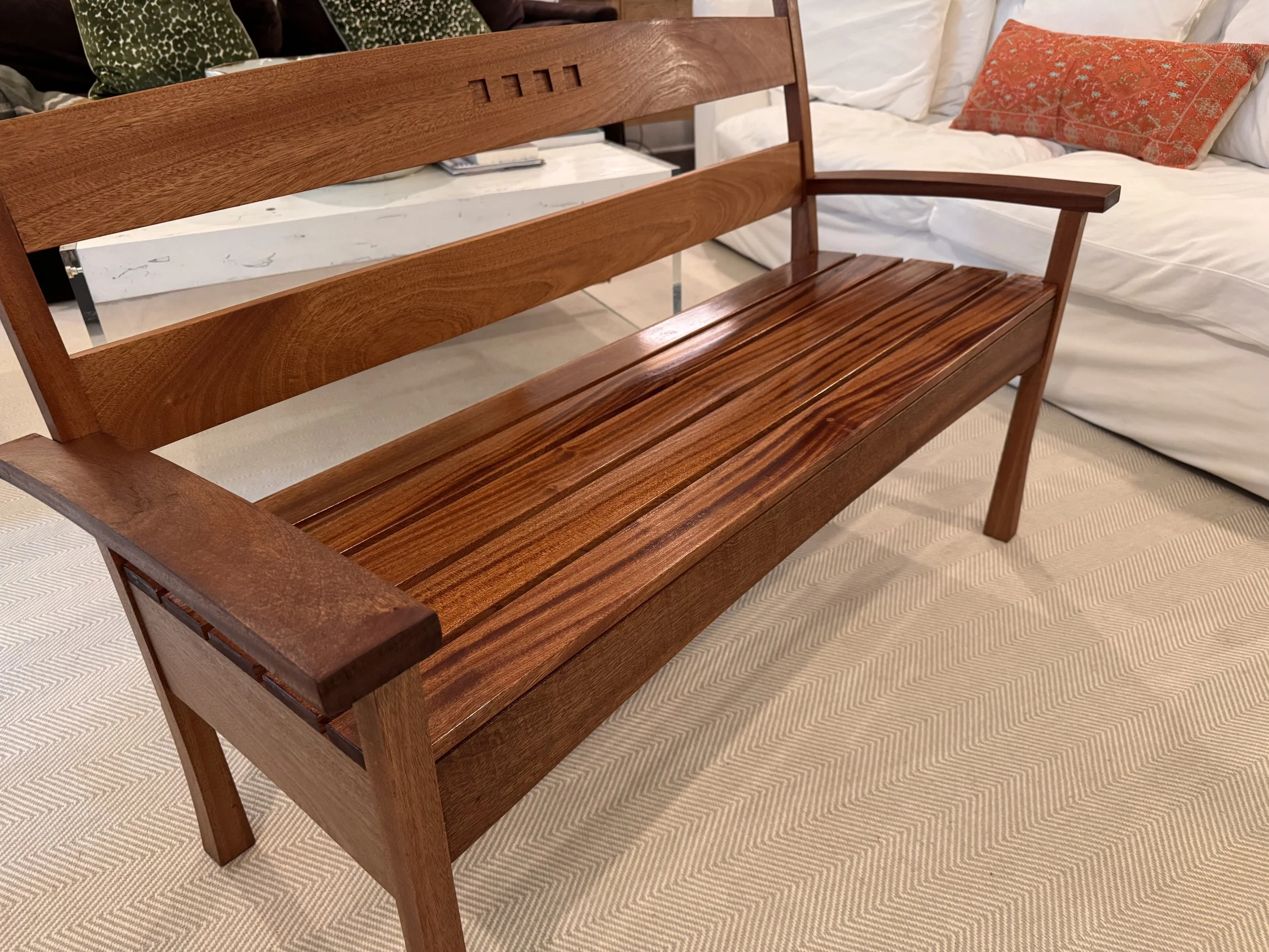 A wooden bench with a slatted seat and backrest, placed on a beige carpeted floor in a living room. In the background, there is a white couch with a decorative orange and patterned pillow, and a coffee table with a white marble top.