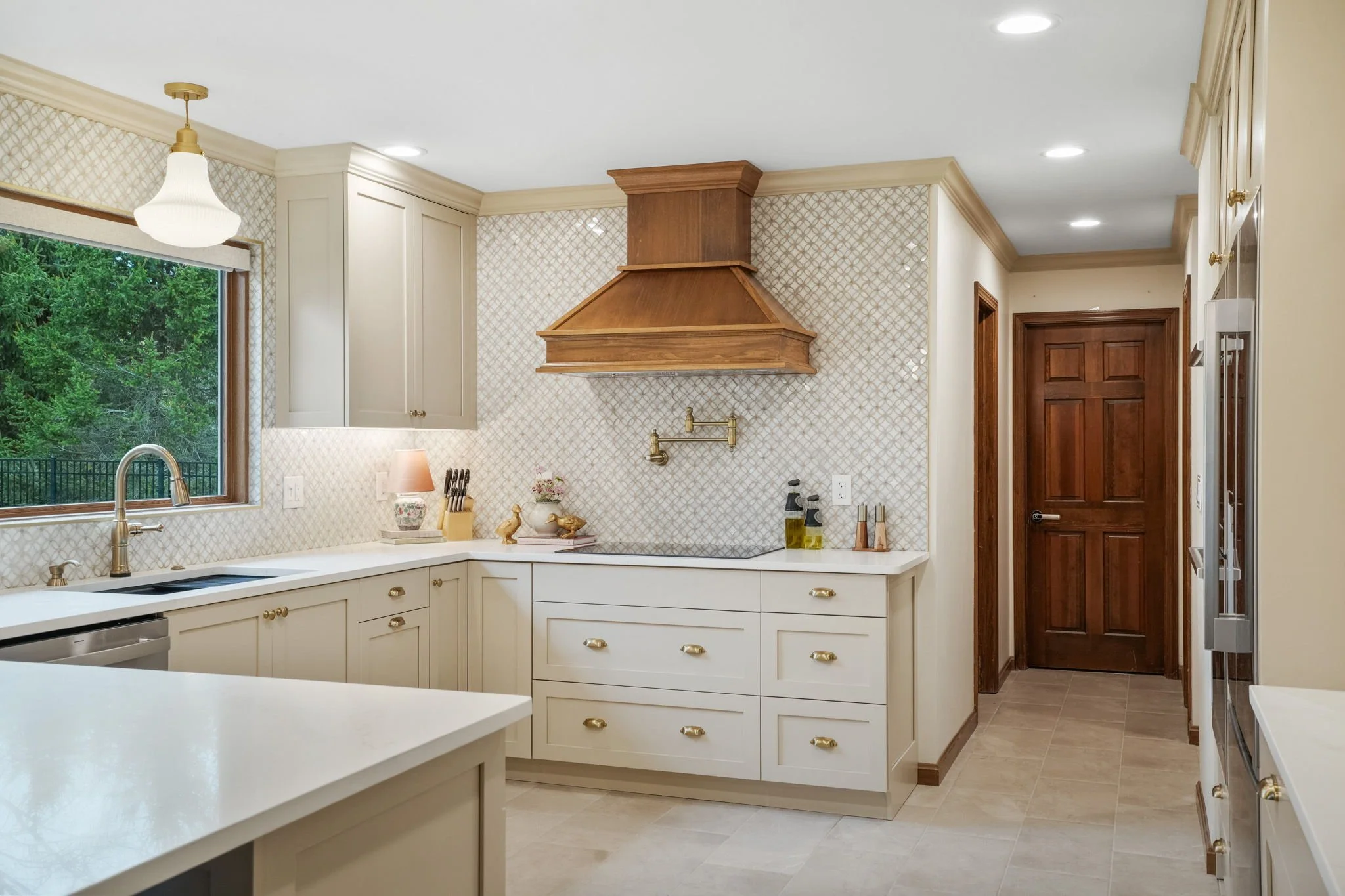 Bright kitchen with cream-colored cabinets, gold hardware, a window overlooking greenery, a wooden range hood, and various kitchen accessories.