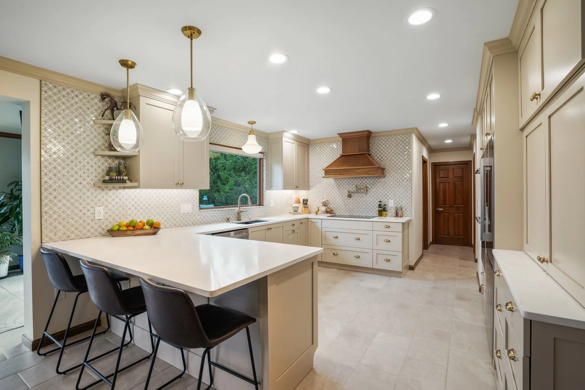 Bright kitchen with white cabinets, beige backsplash, and a central island with black chairs, a window above the sink, and wooden accents on the range hood and door.