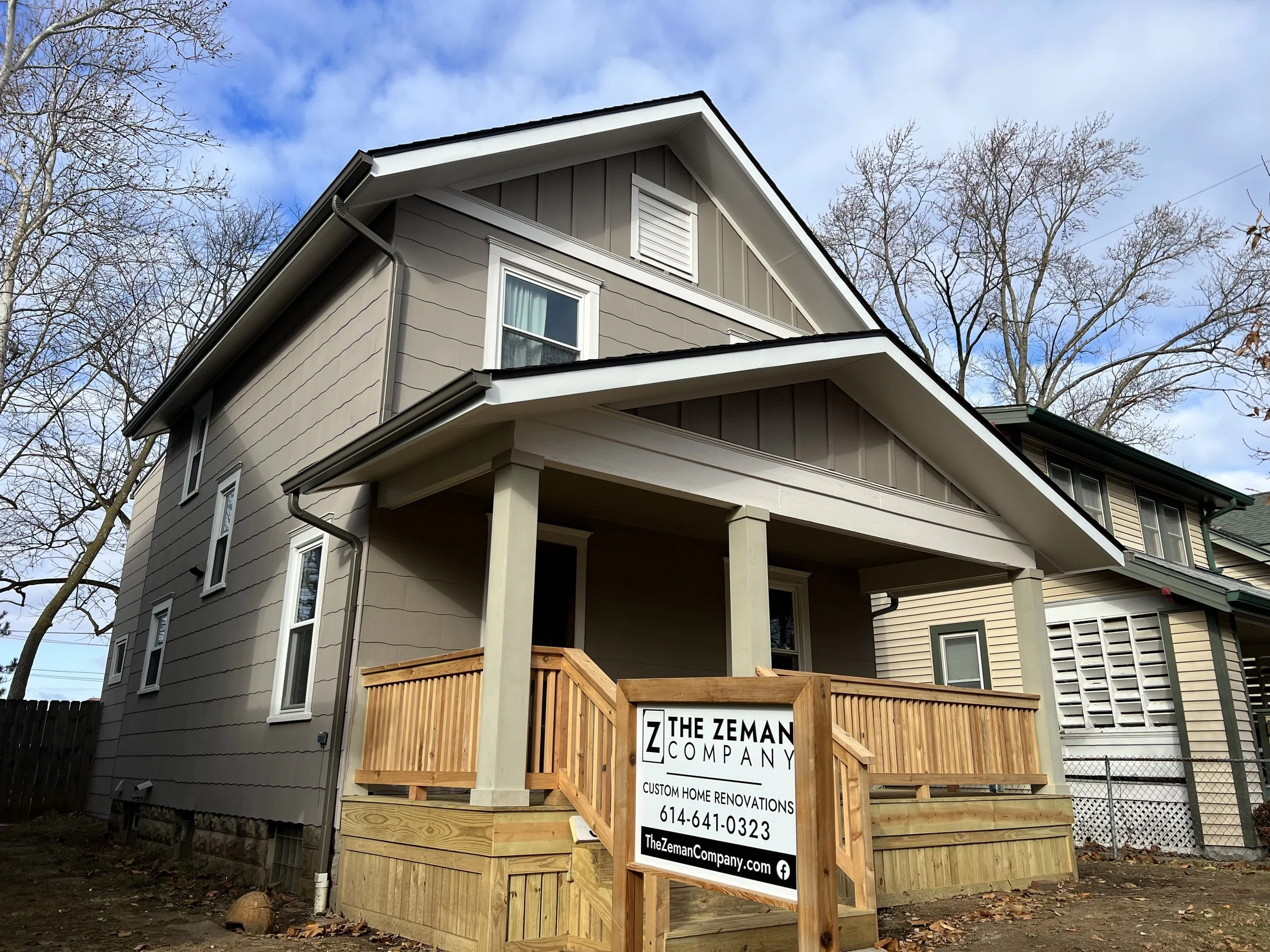 Newly renovated house with a wooden front porch, a sign for The Zeman Company, a home renovation business, in front, and neighboring homes in the background, under a partly cloudy sky.