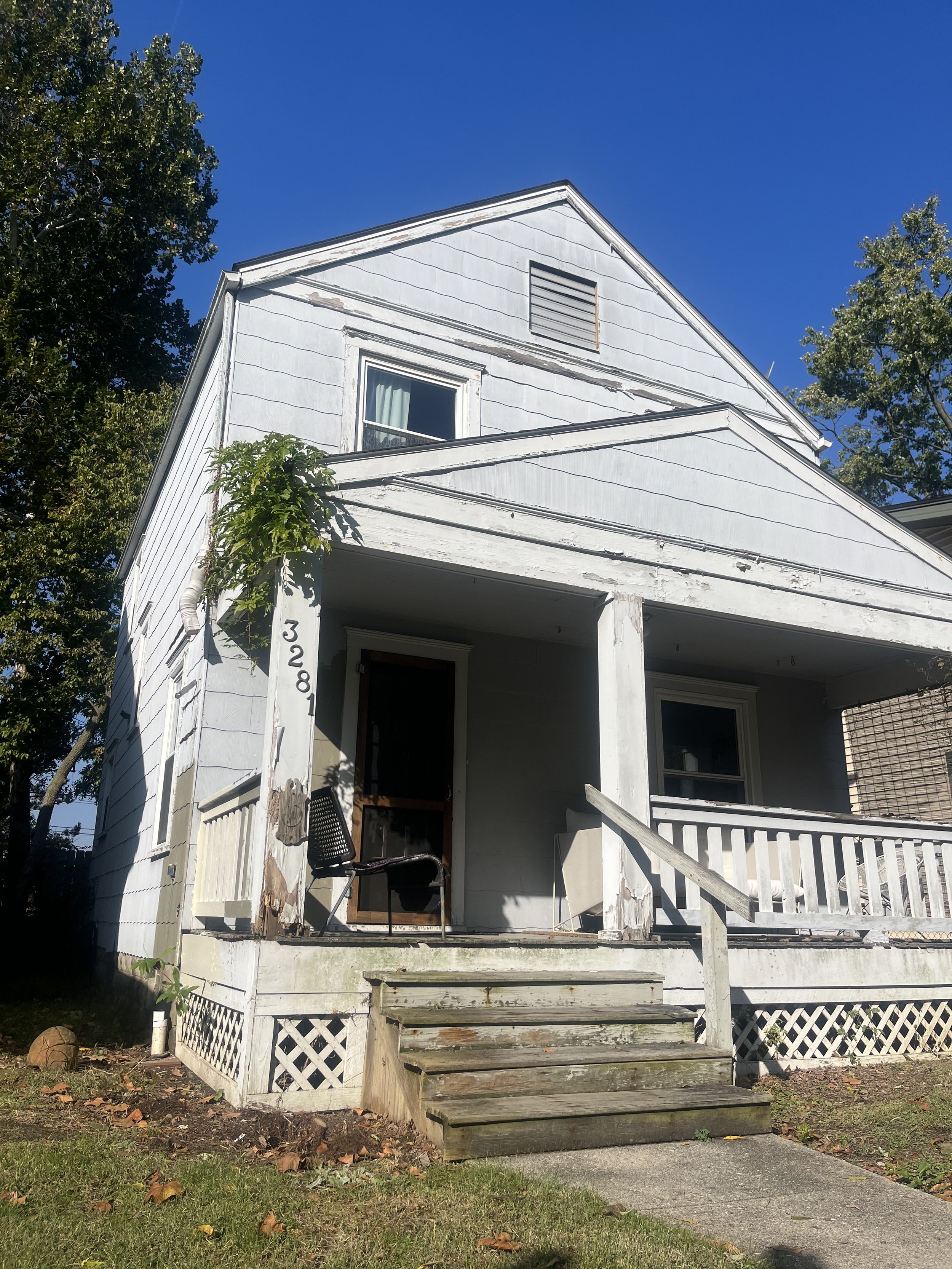 A two-story white wooden house with a front porch, wooden stairs, and a black chair. The house has peeling paint and visible signs of wear. There are trees on each side and a clear blue sky overhead.