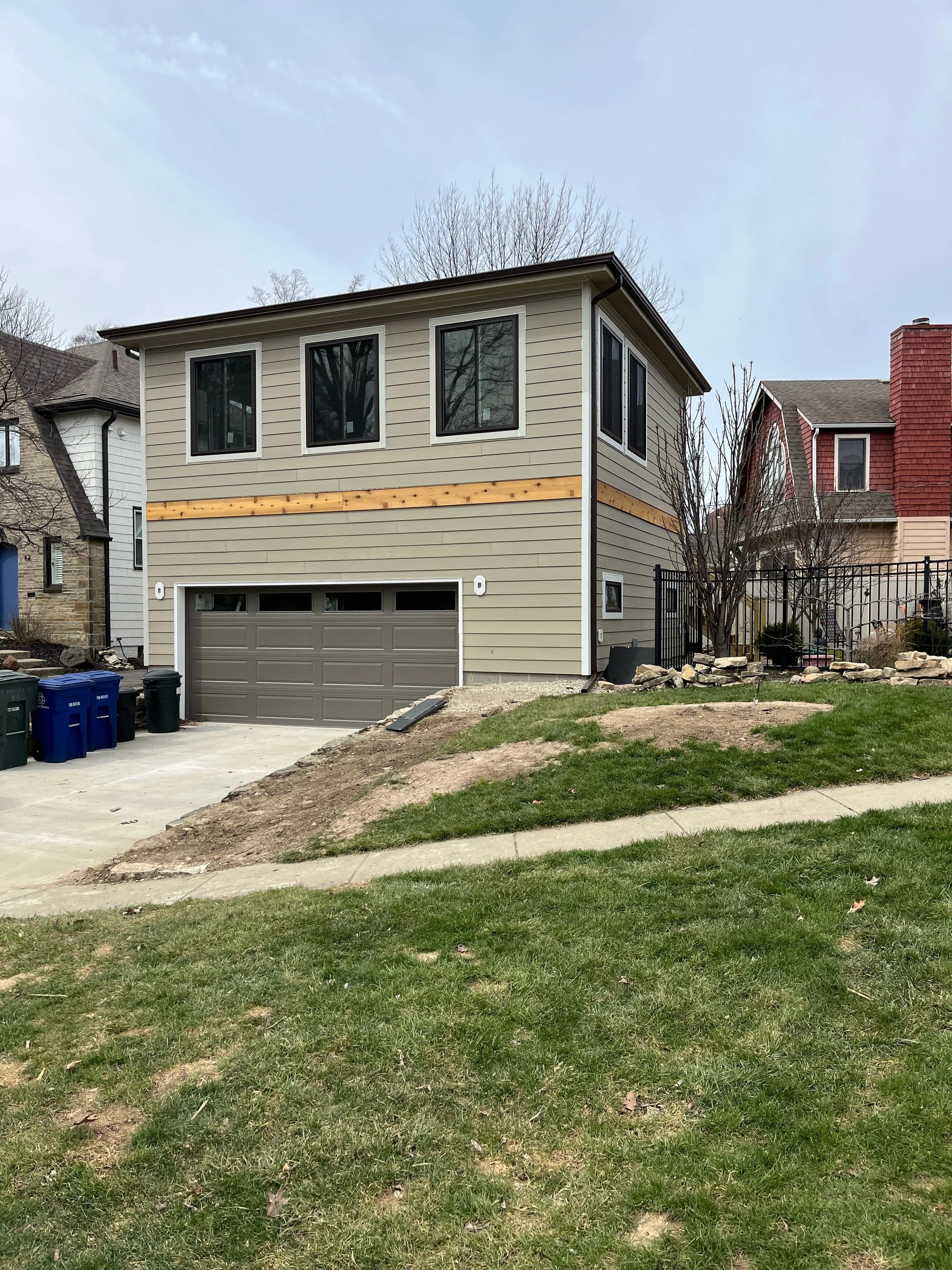 A two-story house under construction with beige siding, three windows on the upper floor, and a gray garage door. The front yard has a grassy lawn with a dirt patch, and a driveway with three trash bins nearby. Trees and neighboring houses are visibl