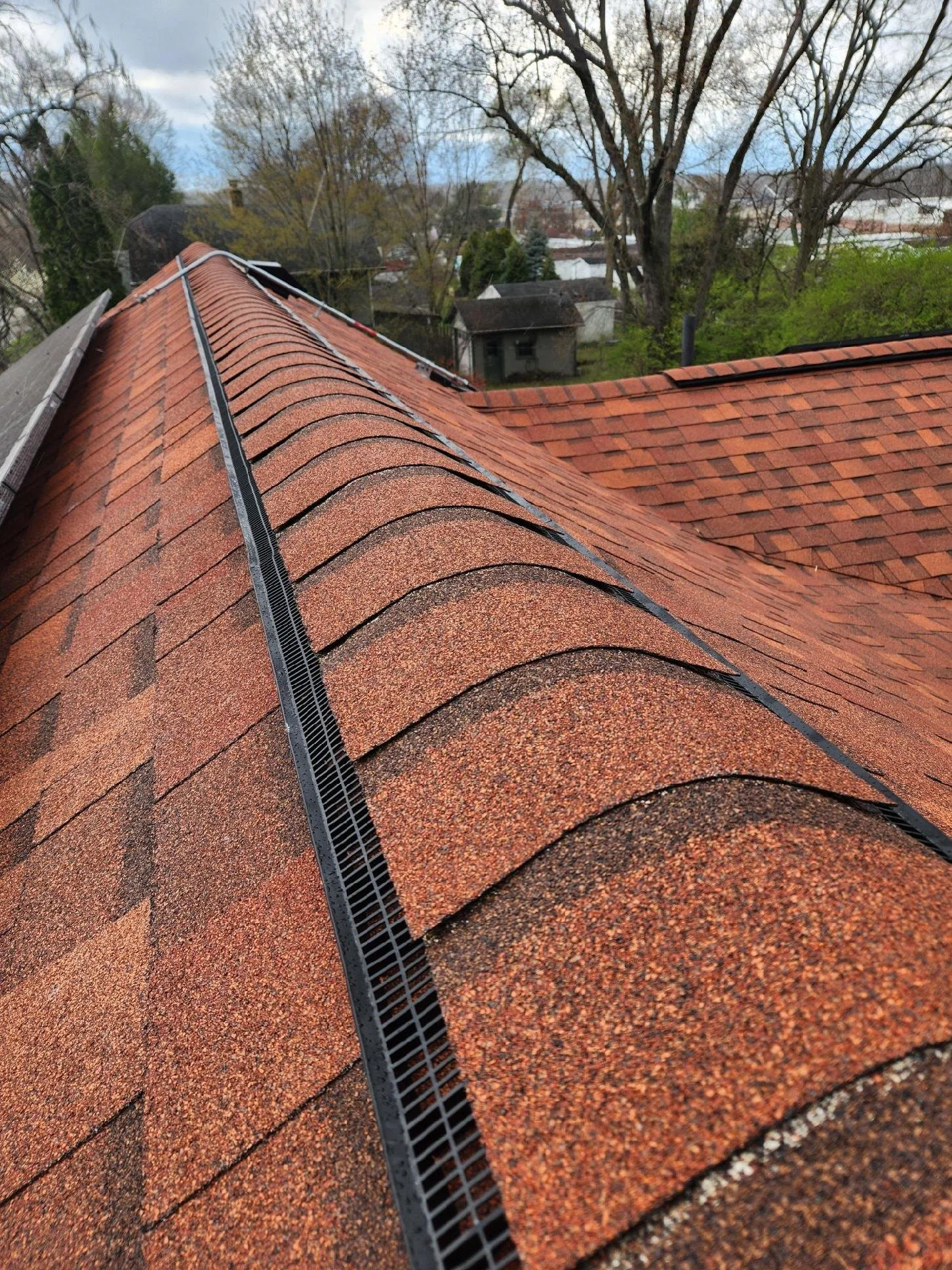 Close-up view of a red shingle roof with a black roof ventilation ridge vent and curved shingles, with trees and other rooftops in the background.