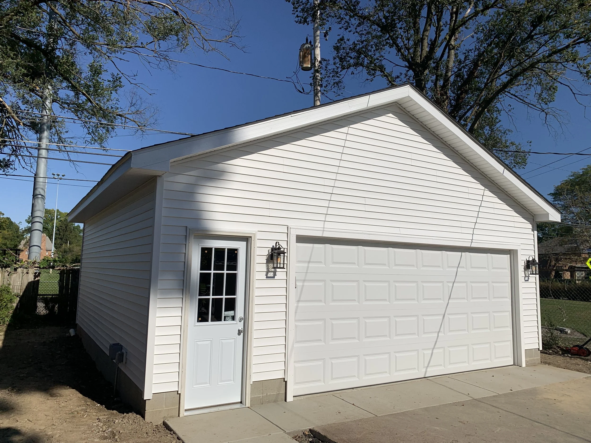 A white garage with a side door and two black wall-mounted lamps on either side, situated on a concrete driveway, with a large tree in the background and a clear blue sky.