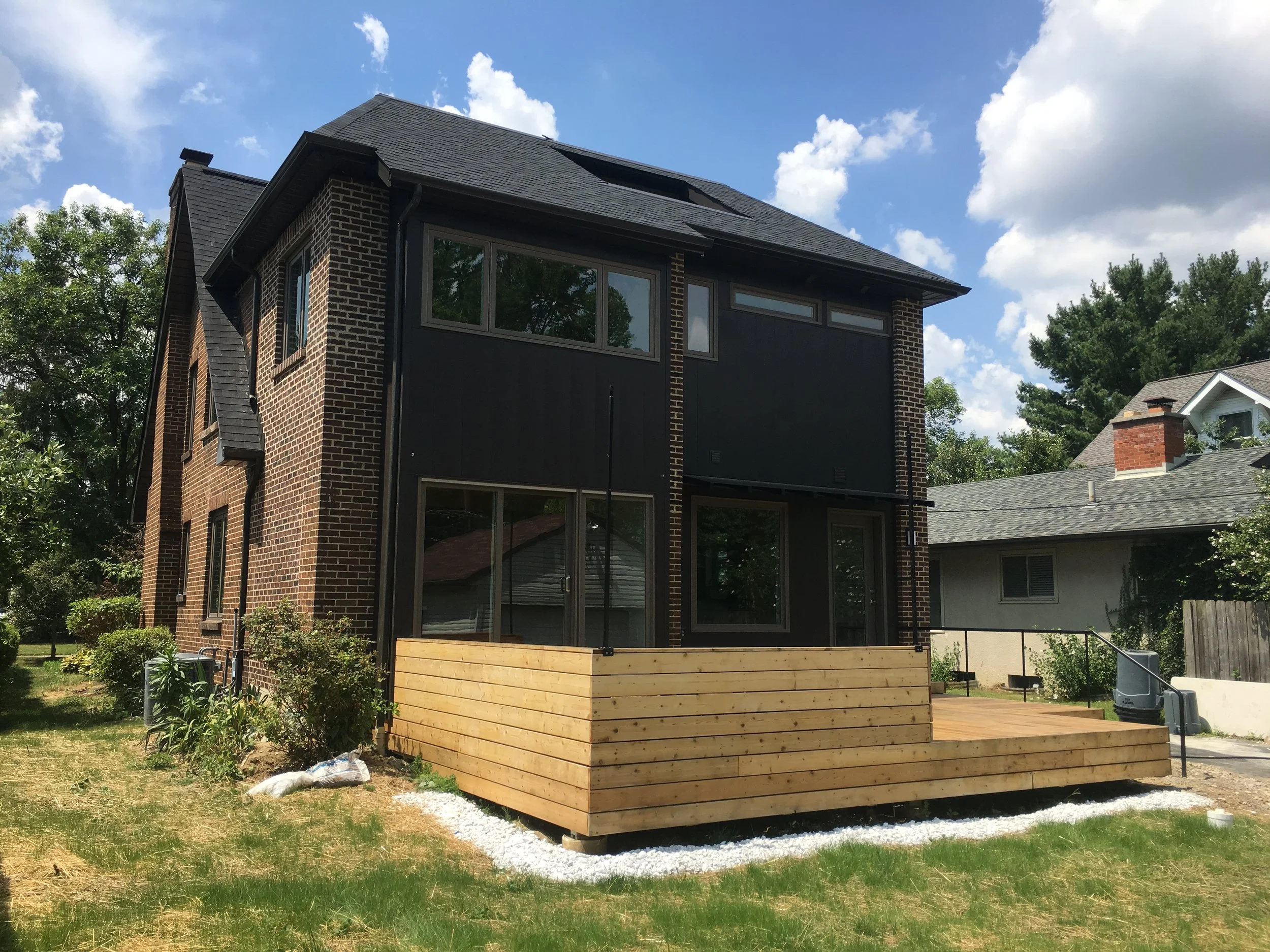 Backyard of a brick house with a partially constructed wooden deck, surrounded by grass, bushes, and neighboring houses, under a blue sky with white clouds.
