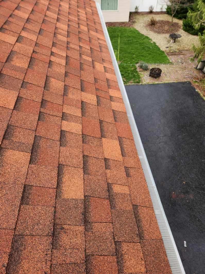 Close-up view of a roof with red asphalt shingles, with part of a backyard visible below.