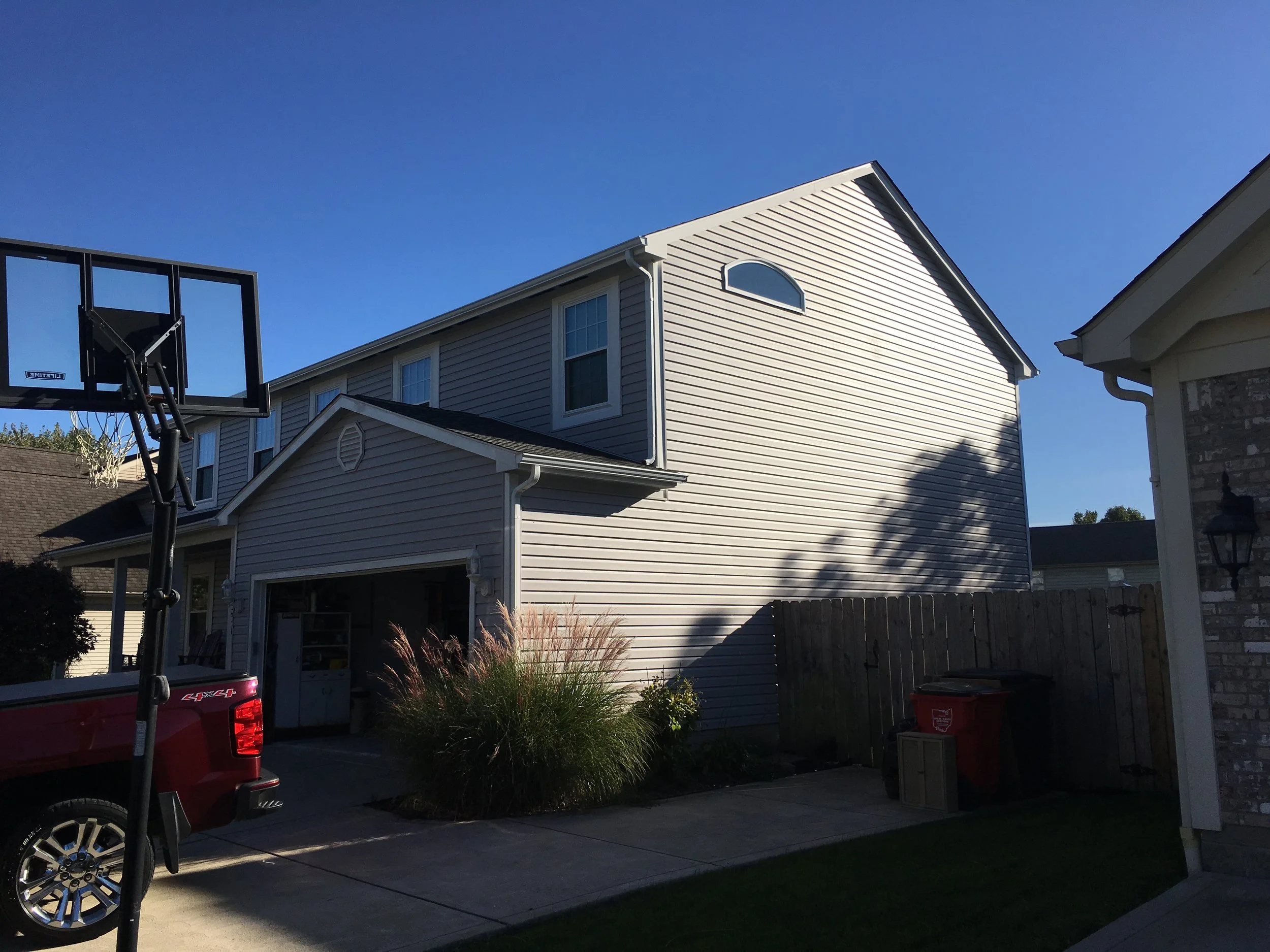 A two-story house with vinyl siding, a garage open showing storage, a basketball hoop, and a small yard with plants and trash bins.