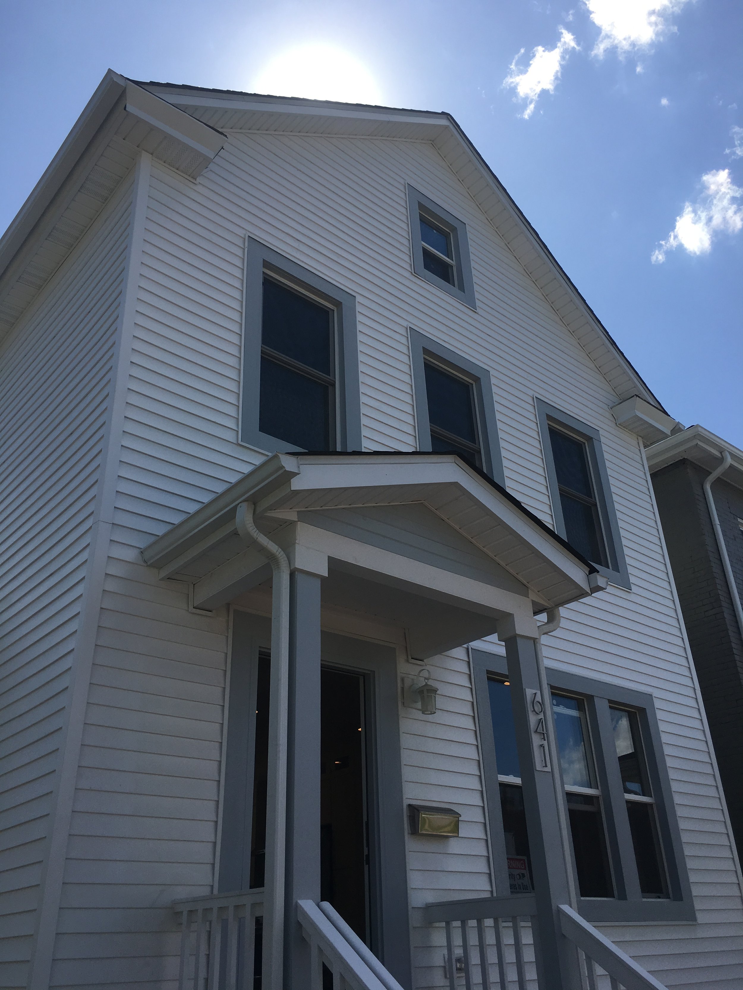 White three-story house with gray trim, porch, and stairs, under a bright sun and blue sky with clouds.