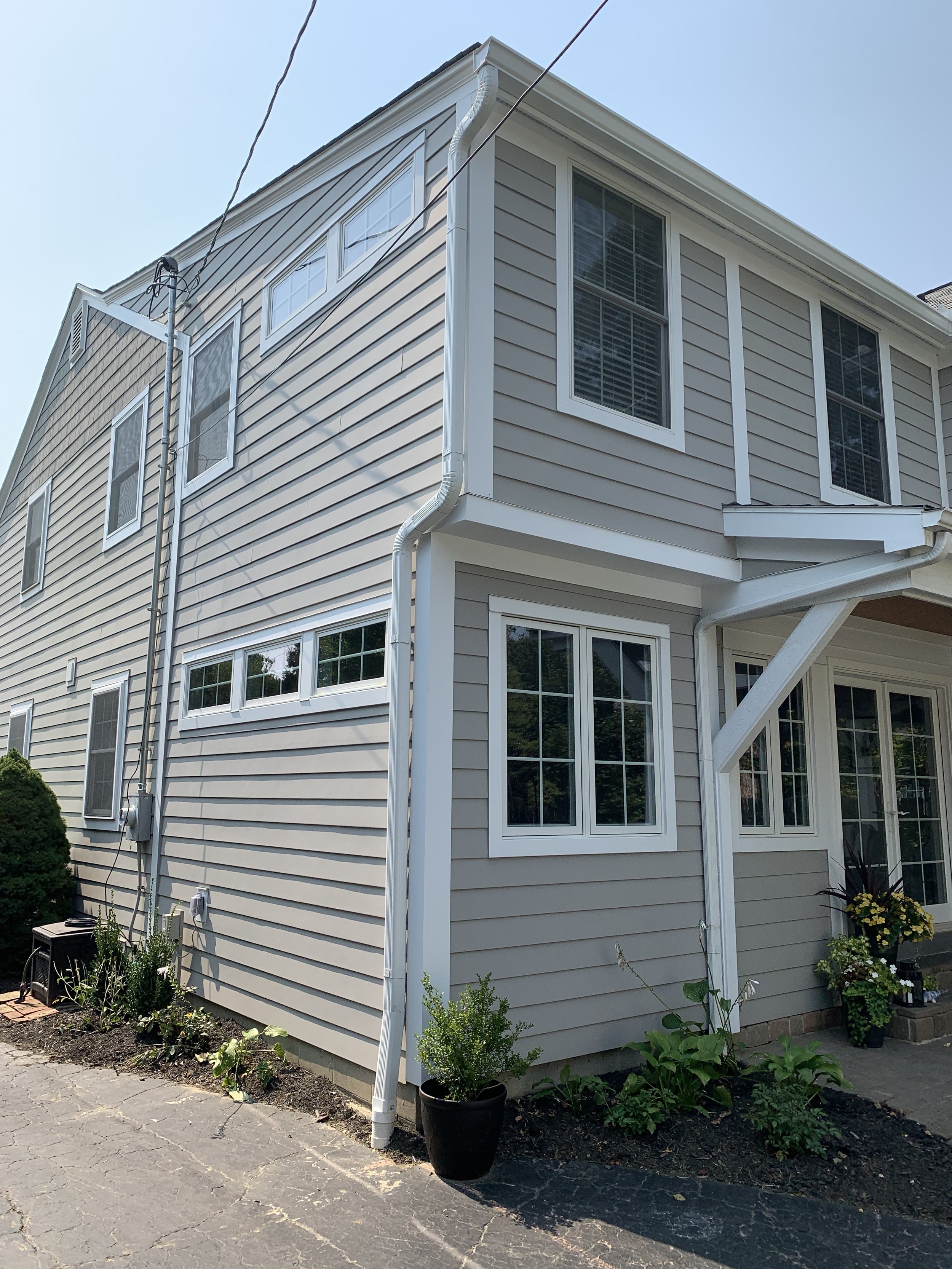 Exterior view of a beige two-story house with white window frames and lattice, surrounded by small plants and a paved driveway.