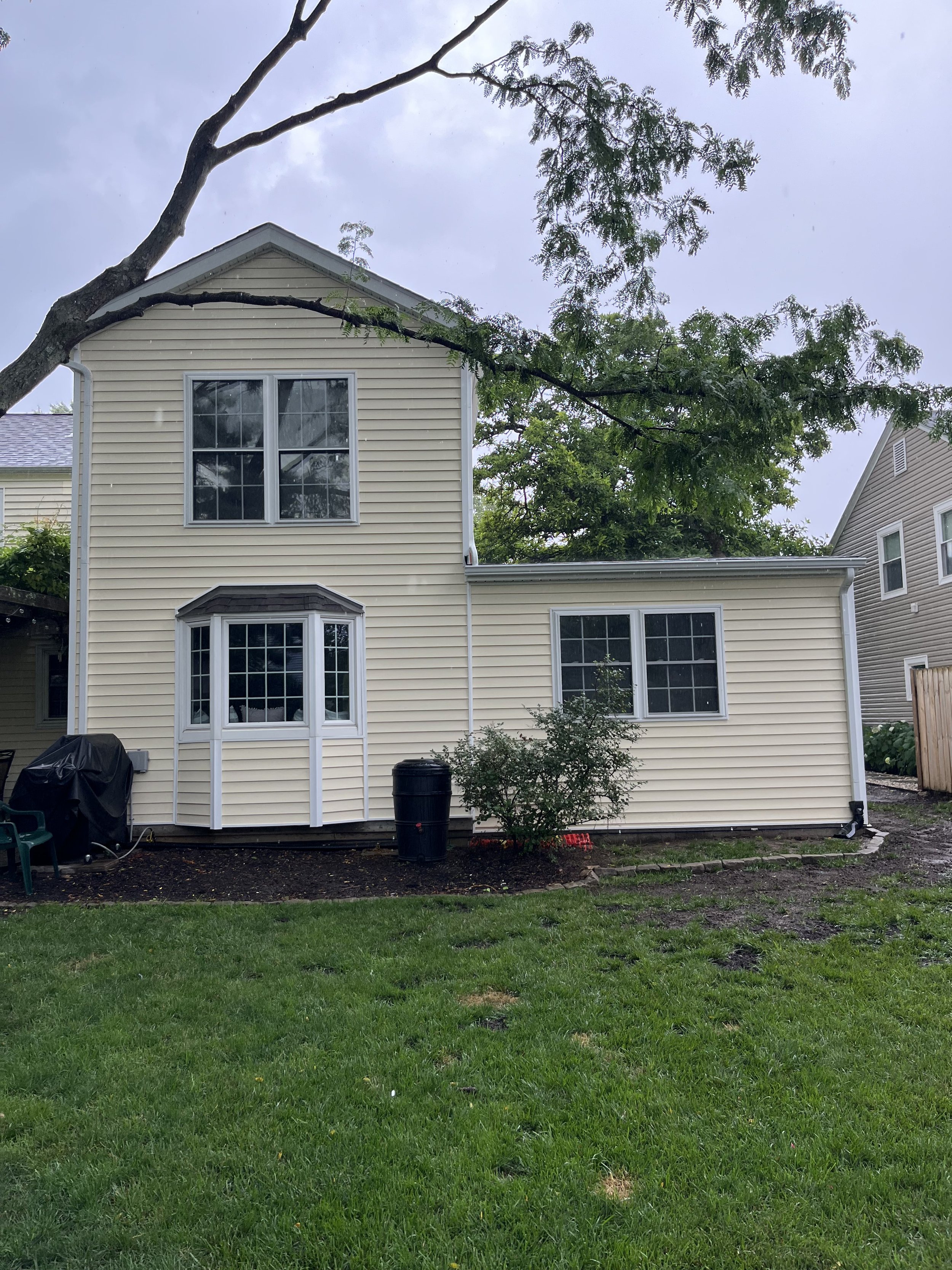 Front view of a beige two-story house with four windows, a bay window on the first floor, and a small tree in the yard surrounded by lawn, mulch, and other vegetation.