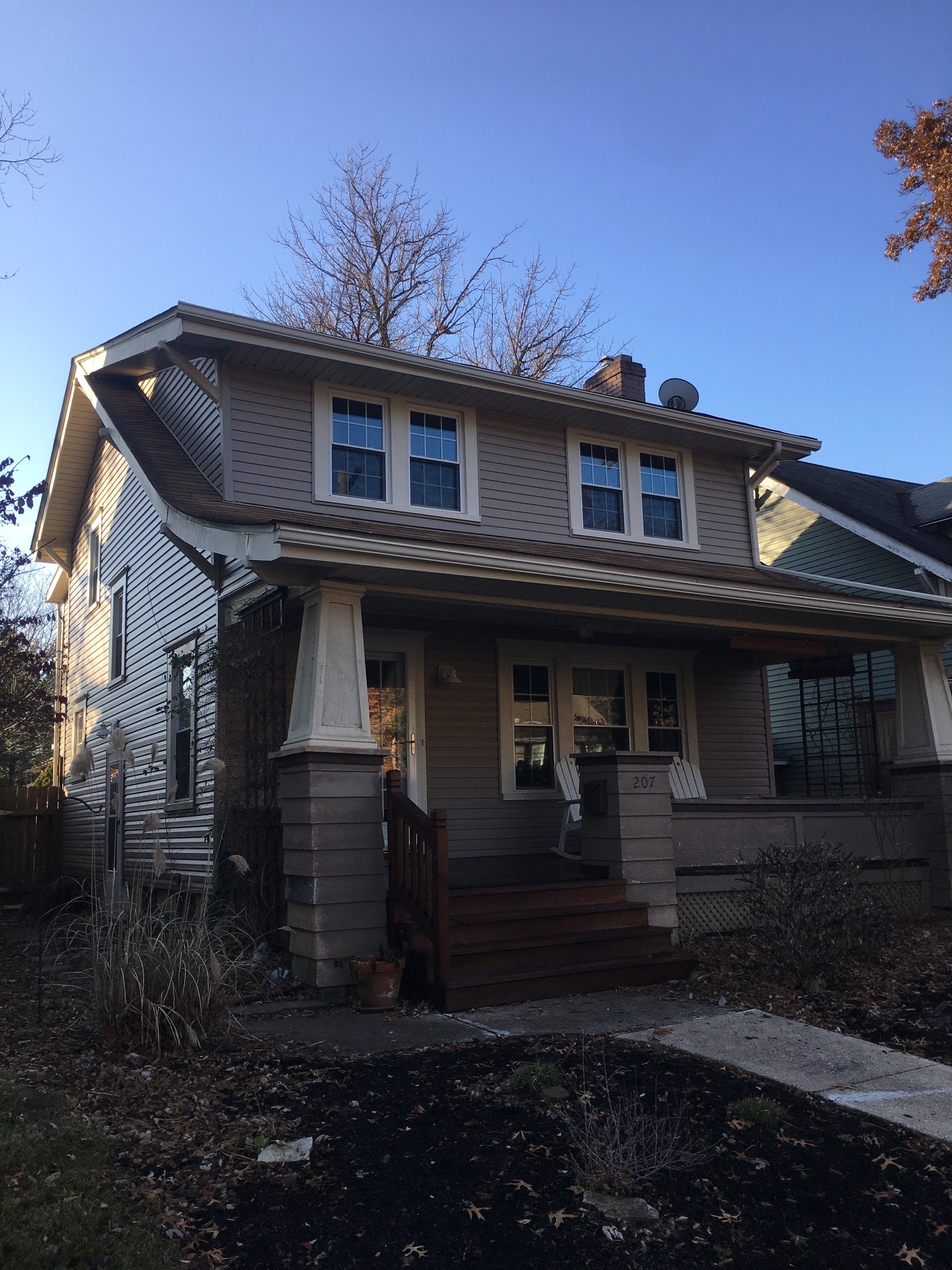 Front view of a two-story house with beige siding, white trim, and a front porch with wooden steps, black metal railings, and a white rocking chair. There are three windows on the upper floor and several on the lower floor. The yard has some plants a