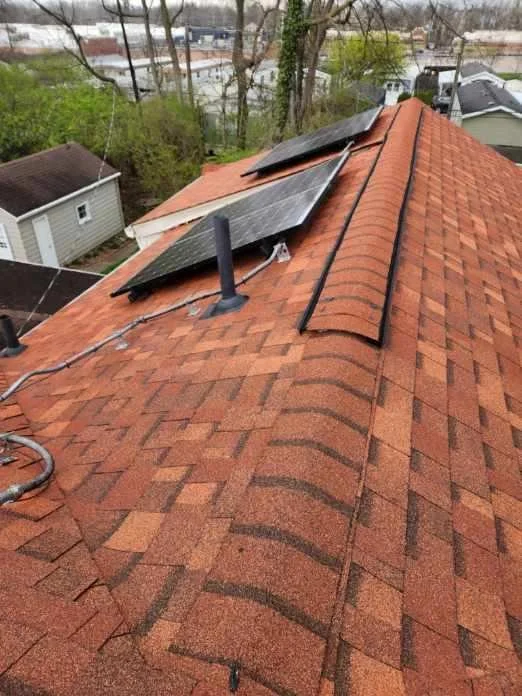 View of a rooftop with three solar panels mounted on reddish-brown shingles, and a vent pipe, in a residential neighborhood with trees and houses in the background.