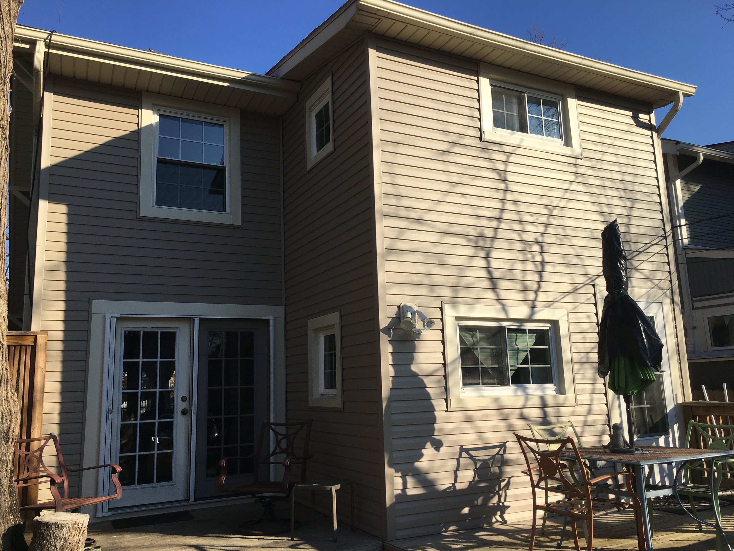 Backyard patio of a beige two-story house with windows, outdoor furniture, a table with an umbrella, and shadows cast by a tree and the house on a clear sunny day.