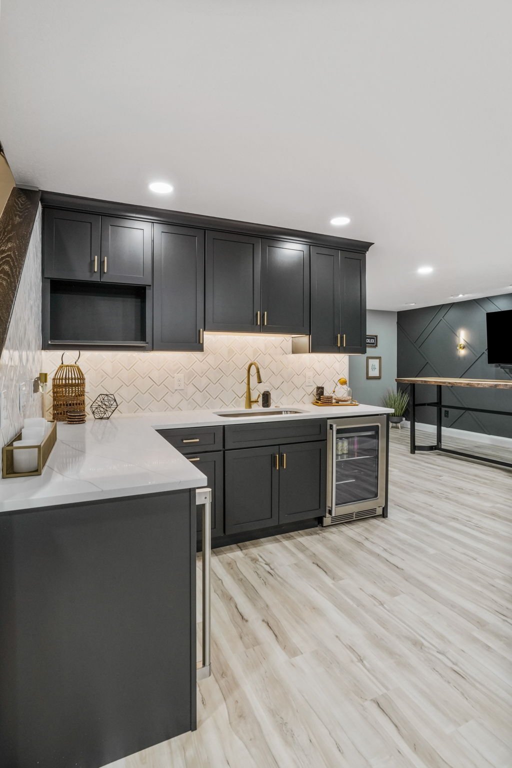 Modern kitchen with dark gray cabinets, white marble countertops, and a white herringbone backsplash. Includes gold fixtures, decorative items, and a small wine fridge, with light wood flooring and a living area visible in the background.