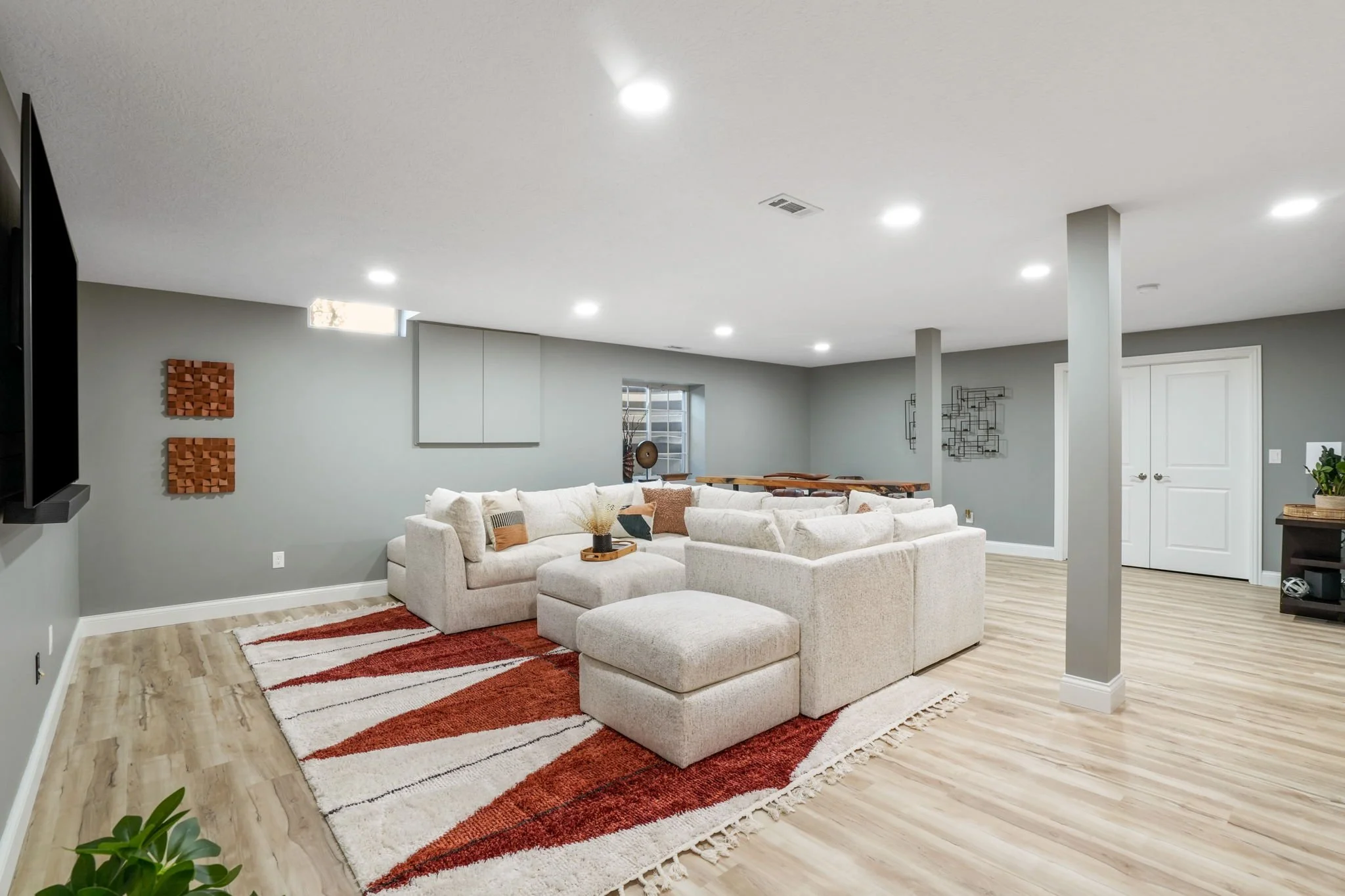 A spacious, modern living room with a large white sectional sofa and pillows, a colorful area rug with orange and cream patterns, gray walls, ceiling lights, a small window with blinds, wall art, and a corner with plants and decor.
