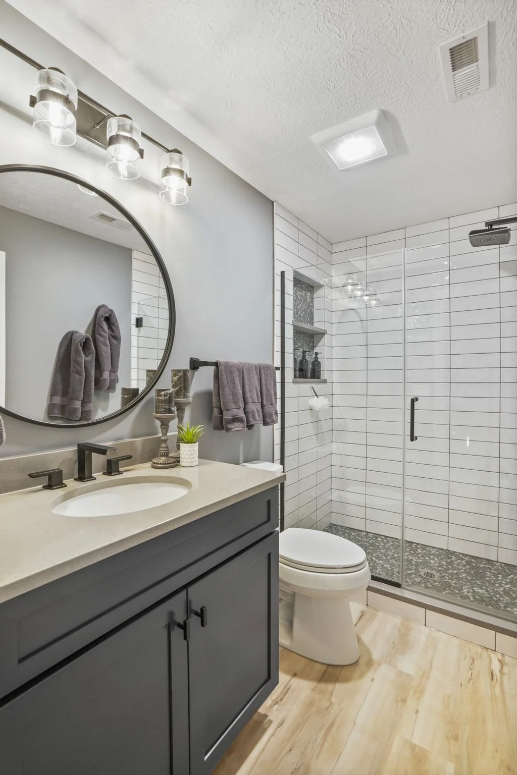 A modern bathroom interior featuring a gray vanity with a beige countertop, a round mirror, gray towels, a small plant, and a shower with a glass door, white subway tiles, and a pebble tile floor.