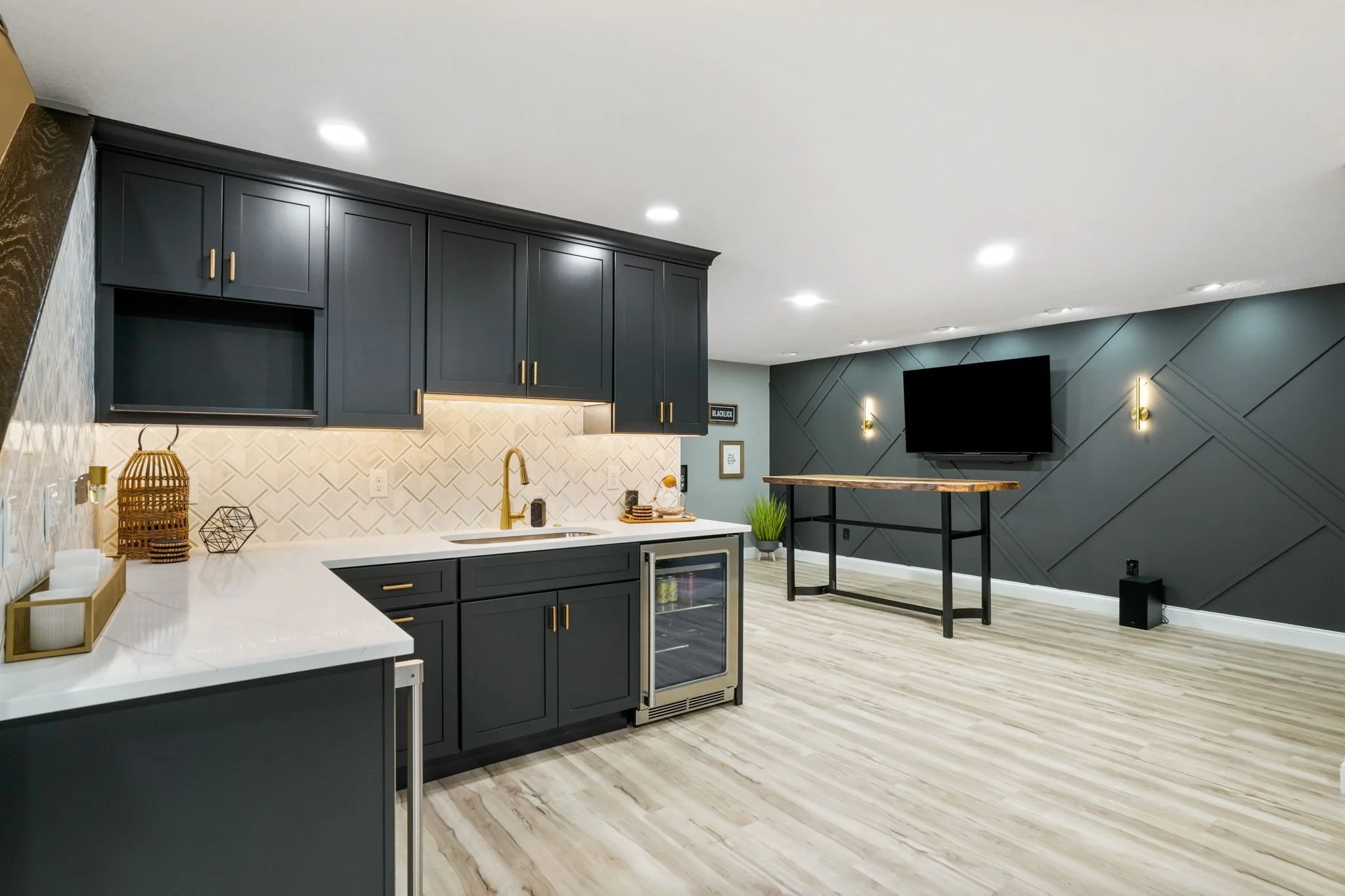 Modern kitchen with dark navy cabinets, white countertops, beige tiled backsplash, small wine fridge, gold accents, and open living space with a TV and dark gray accent wall