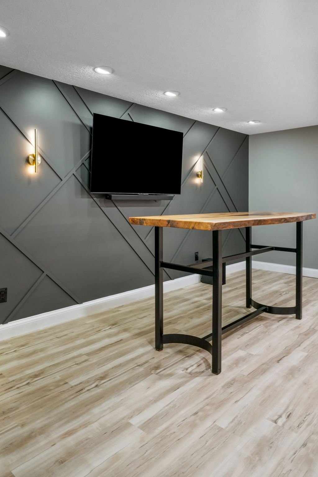 Modern interior with a gray geometric wall, a wall-mounted flat-screen TV, two gold wall sconces, and a wooden table with black metal legs on wood flooring.