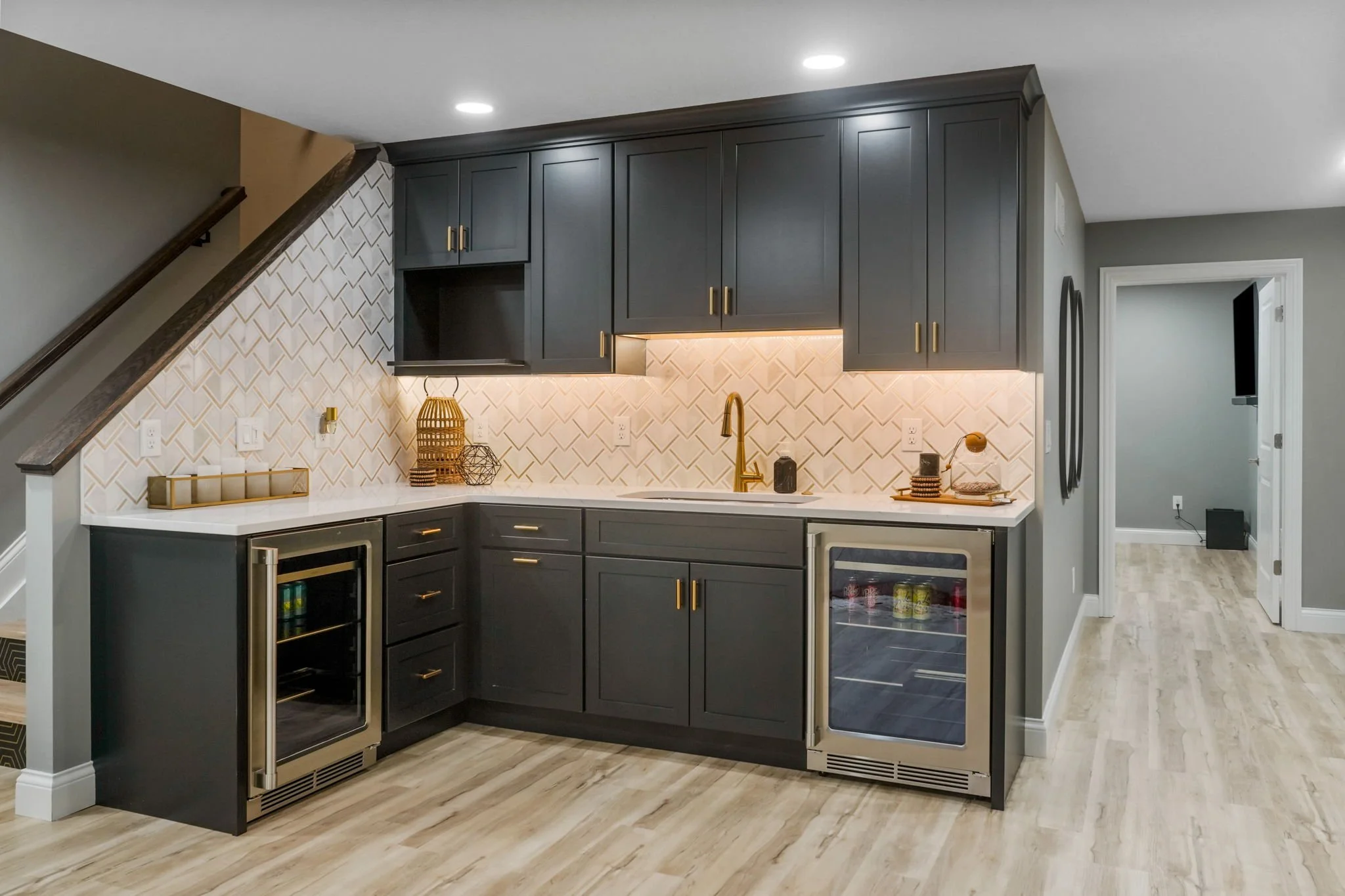 Modern kitchen with dark gray cabinets, a white countertop, and under-cabinet lighting. Small wine cooler on left and mini-fridge on right, with gold hardware and faucet.