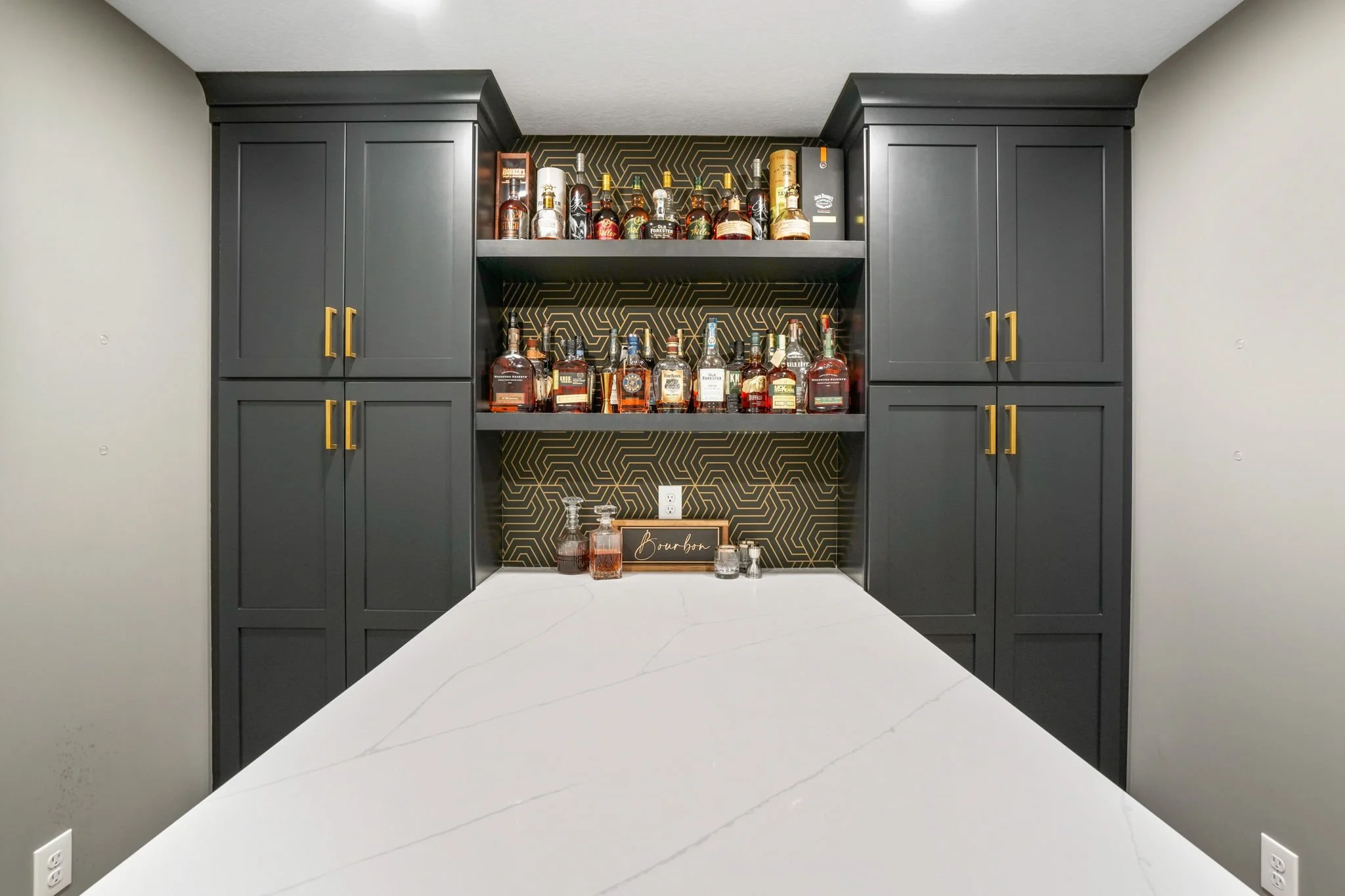 A home bar with black cabinets, gold handles, and a geometric gold and black patterned backsplash. Bottles of various liquors are arranged on shelves in the center.