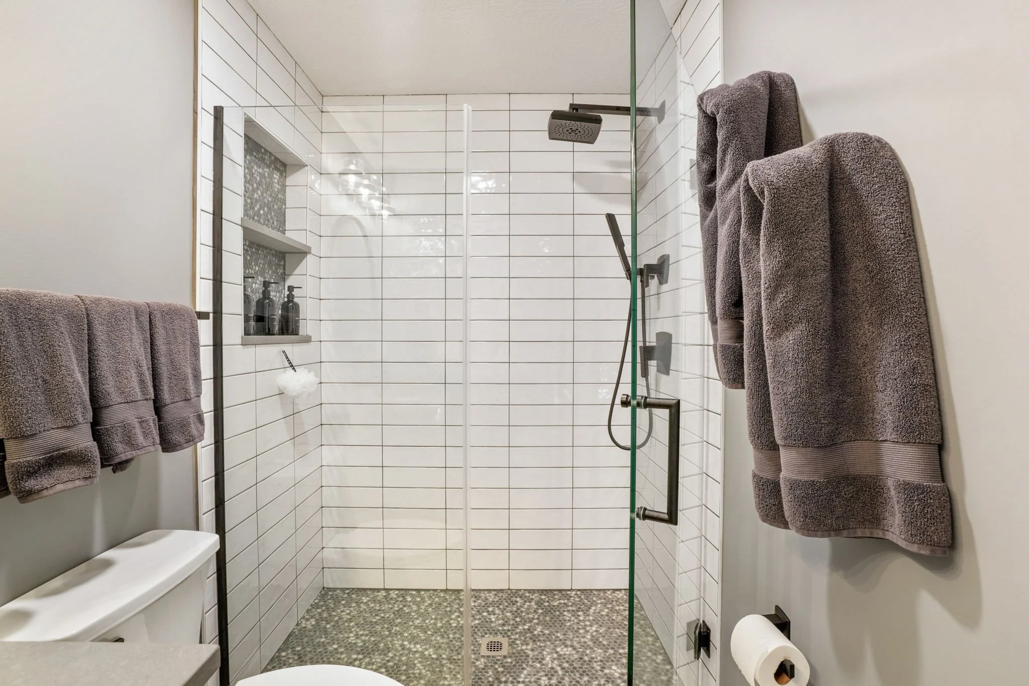 Modern walk-in shower with white subway tiles, pebble tile floor, rain showerhead, and a glass door; grey towels hanging on the wall, and a toilet paper roll visible in the foreground.