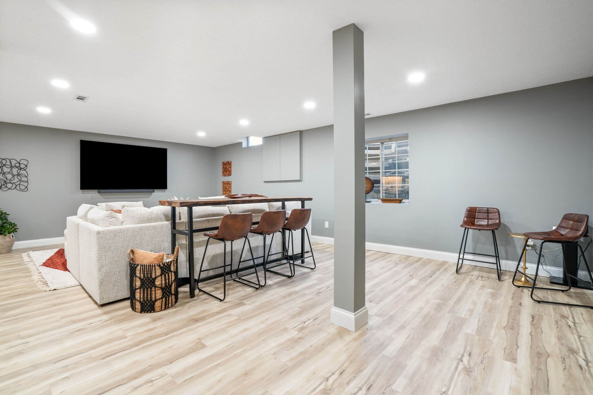 A spacious basement living area with light wood flooring, gray walls, a large white sectional sofa, a wall-mounted flat screen TV, a tall wooden bar table with four brown chairs, a small window, and accent chairs.