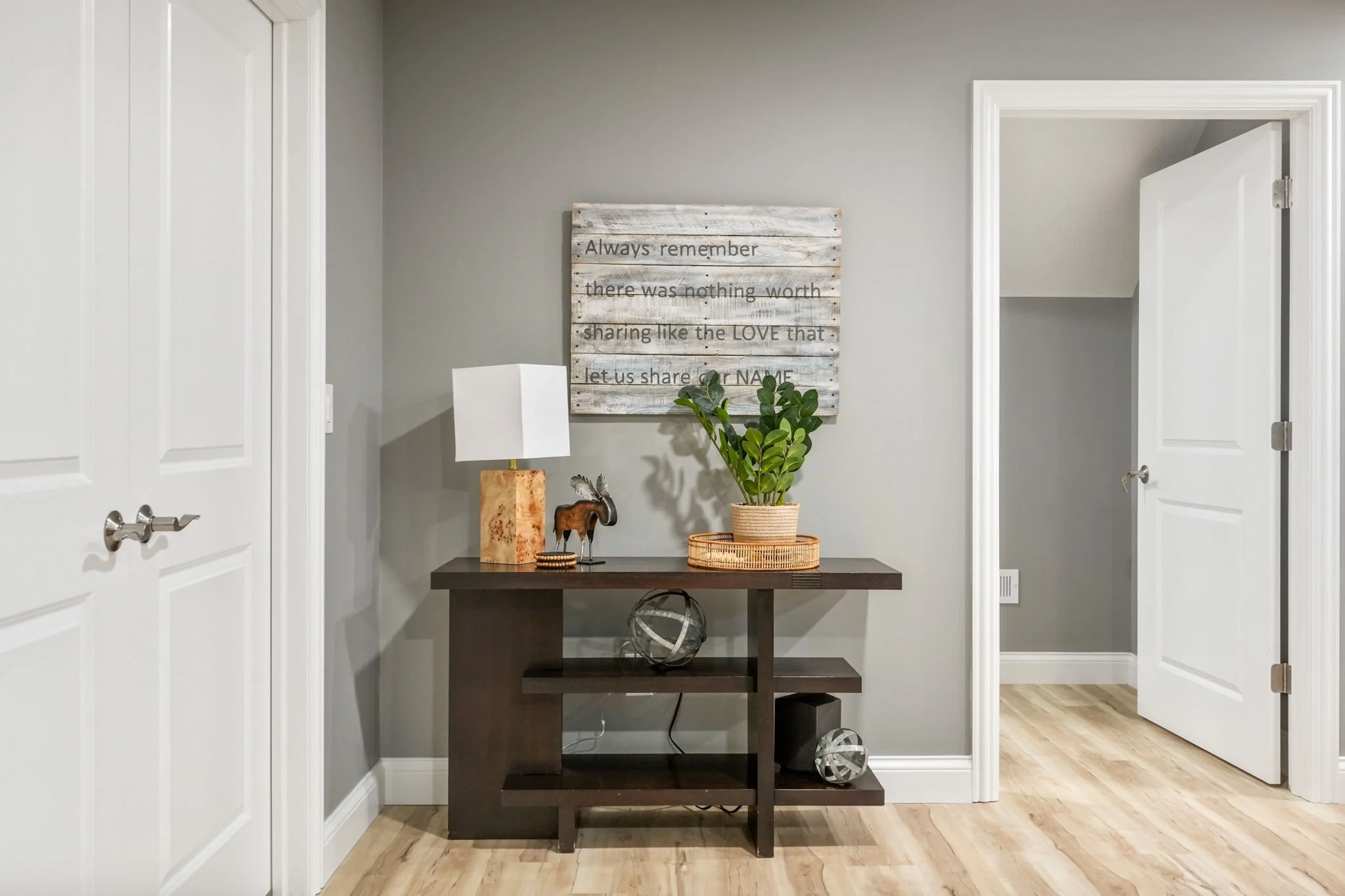 Interior view of a hallway with a dark wooden console table, decorative items including a lamp, a small moose figurine, and a potted plant. A wall art with a sentimental quote hangs above the table, and there are two open white doors on either side.