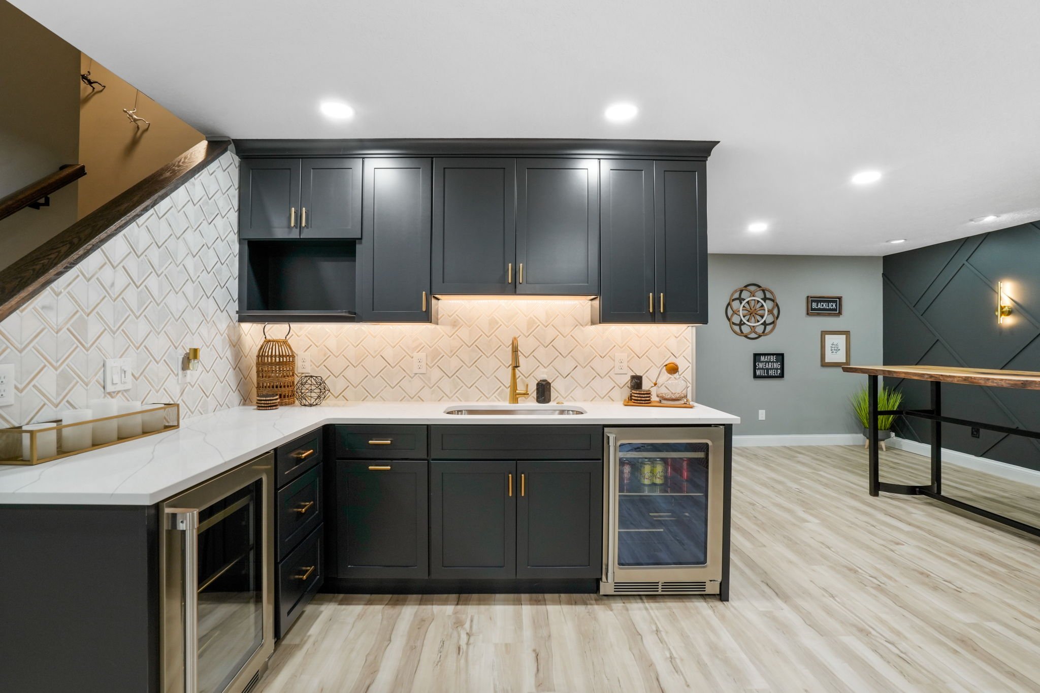 Modern kitchen with dark navy cabinets, white marble countertop, gold hardware, and a herringbone tile backsplash. Contains small appliances, decor, and a staircase nearby.