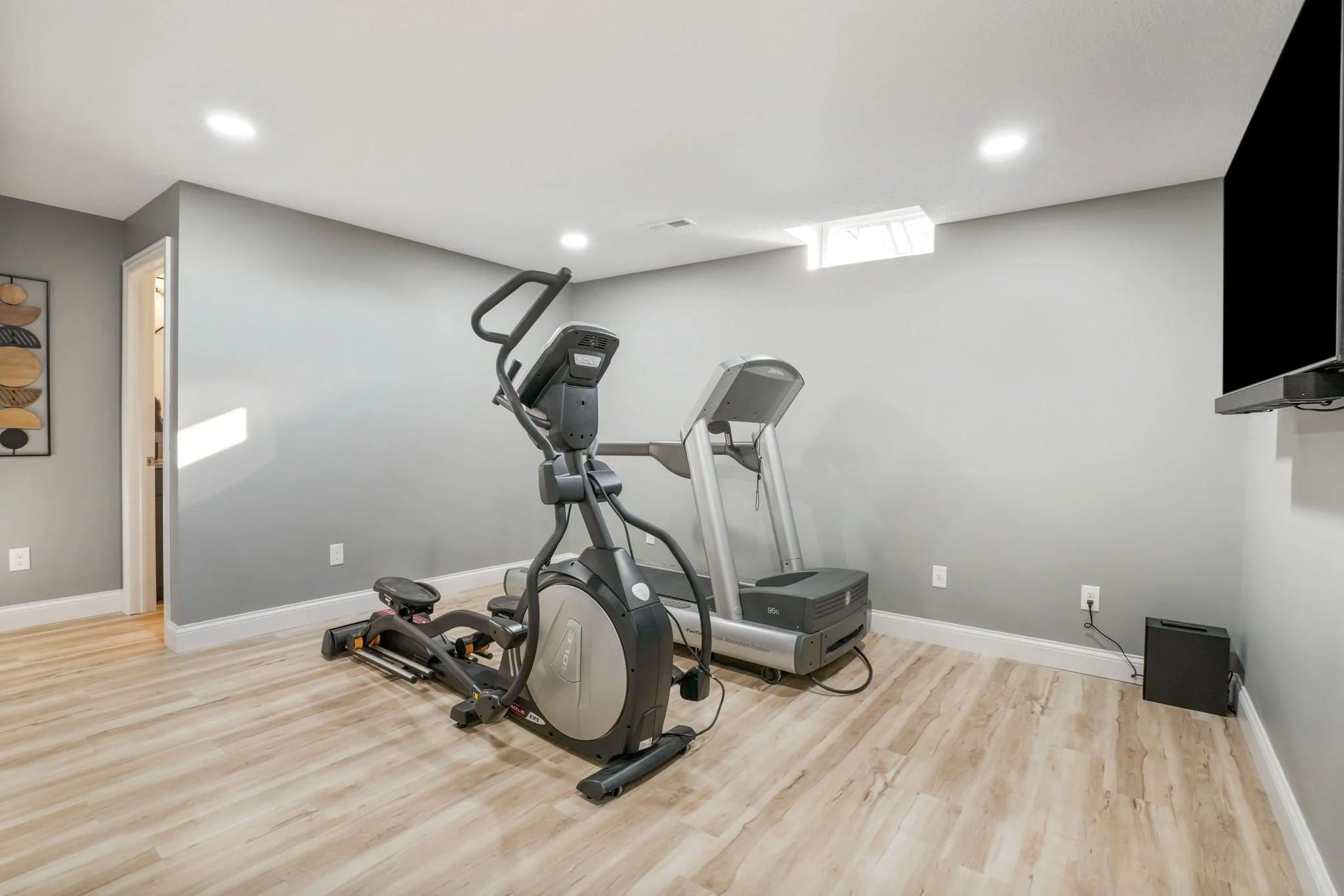 A home gym with light wood flooring, gray walls, and exercise equipment including a recumbent bike and a treadmill in a spacious room.