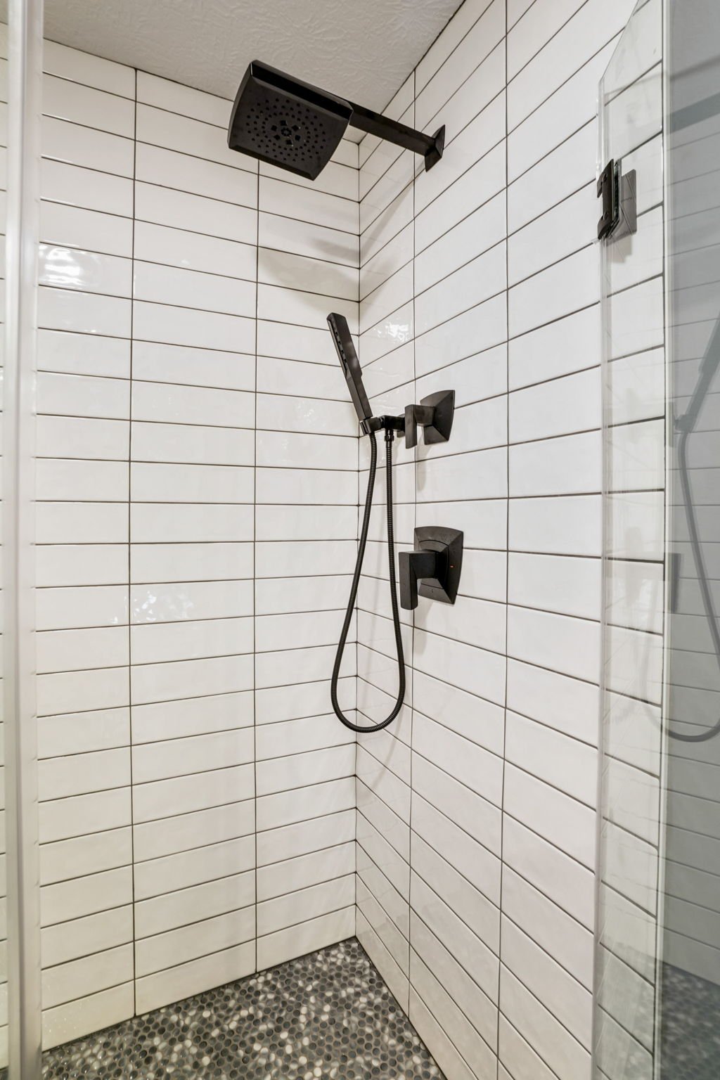 Shower area with white linear tiled wall, black showerhead, handheld shower wand, and pebble stone floor.