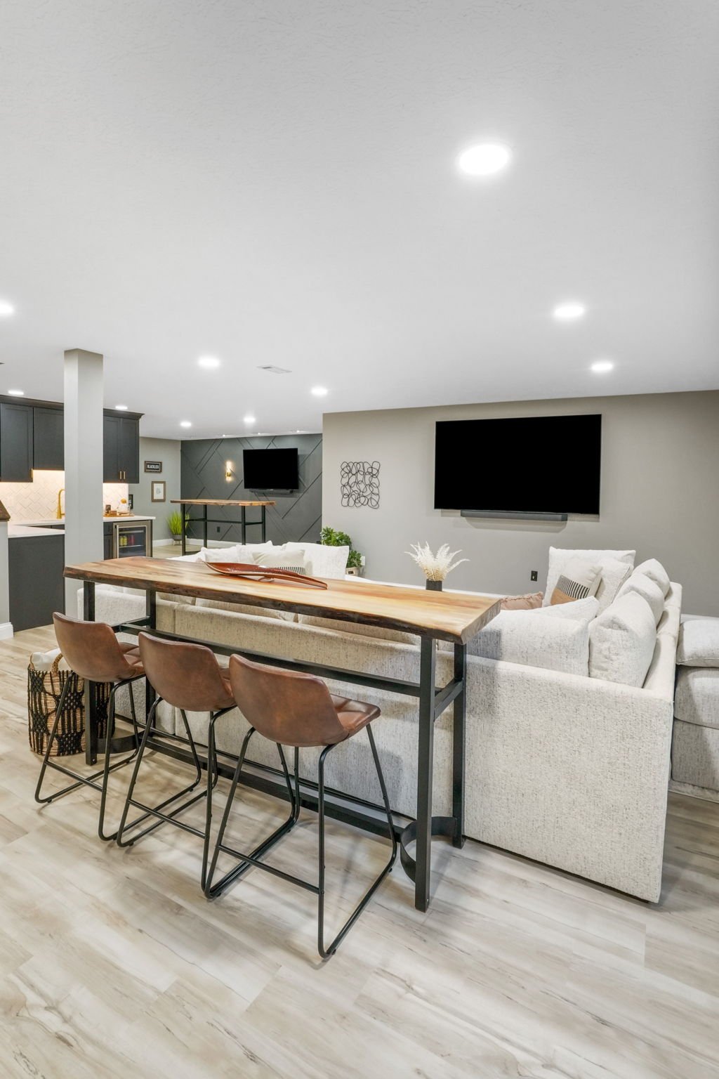Modern living room with white sectional sofa, wooden high-top table with three brown leather chairs, flat-screen TV on gray wall, neutral color palette, and hardwood flooring.