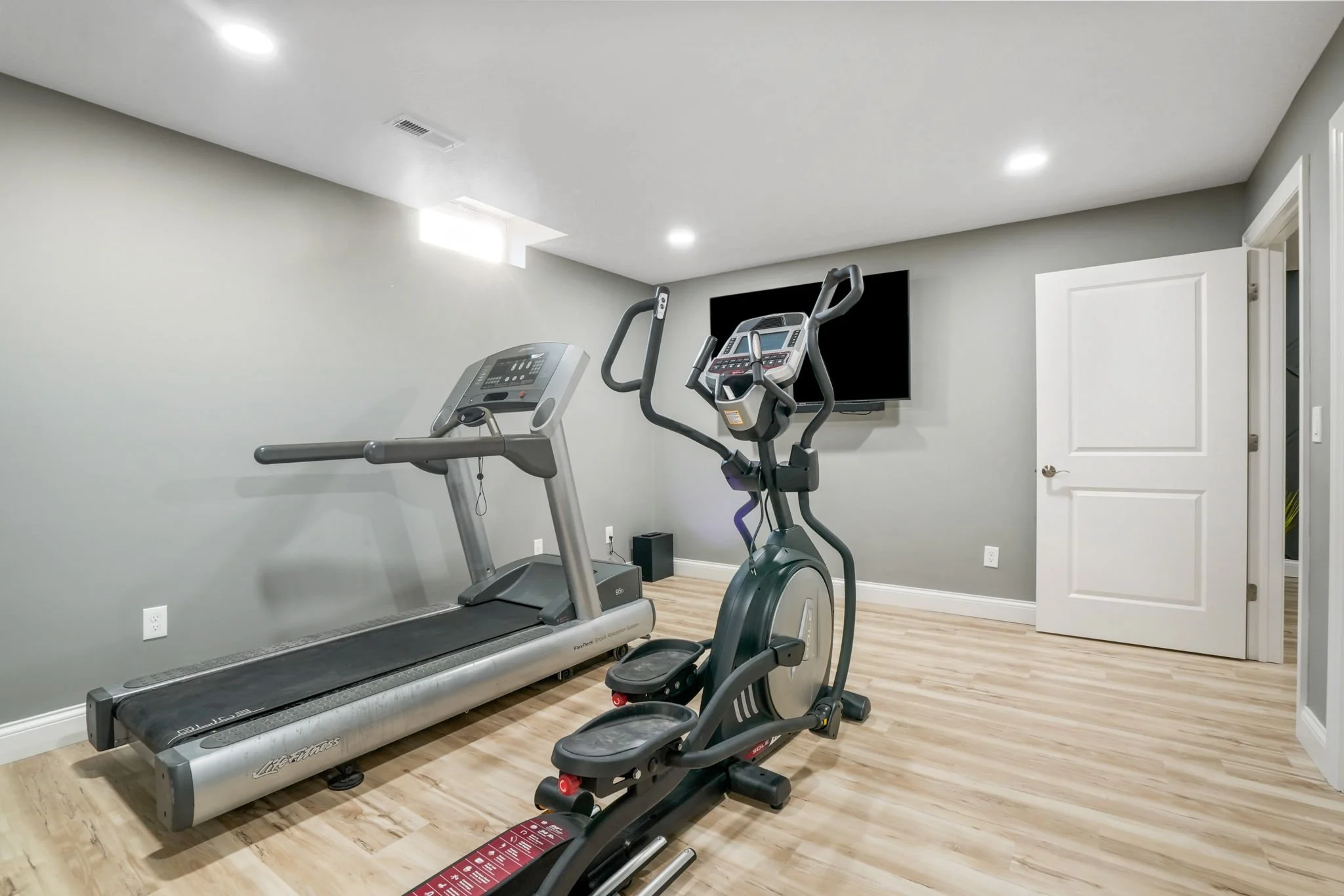 Home gym with treadmill, exercise bike, wall-mounted TV, and wooden floor in a room with recessed ceiling lights.