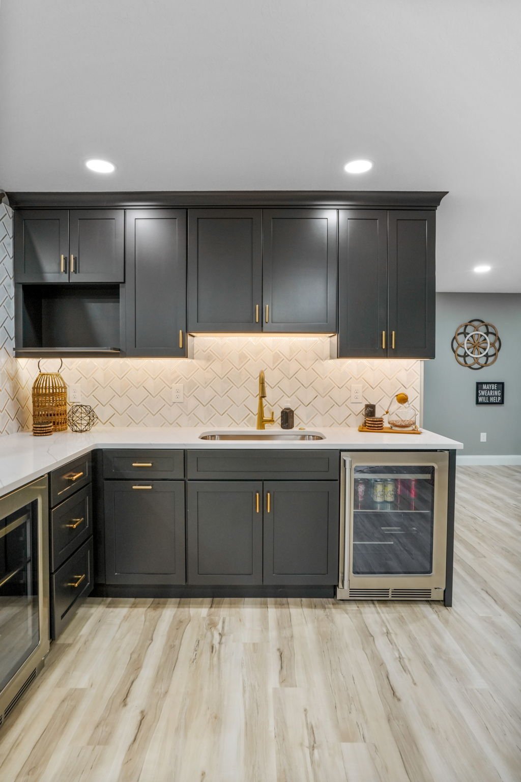 Modern kitchen with dark gray cabinets, white countertops, gold hardware and faucet, backsplash with a diamond pattern, and a small wine cooler at the end of the counter. Decor includes baskets, a decorative sign, and small decorative items.